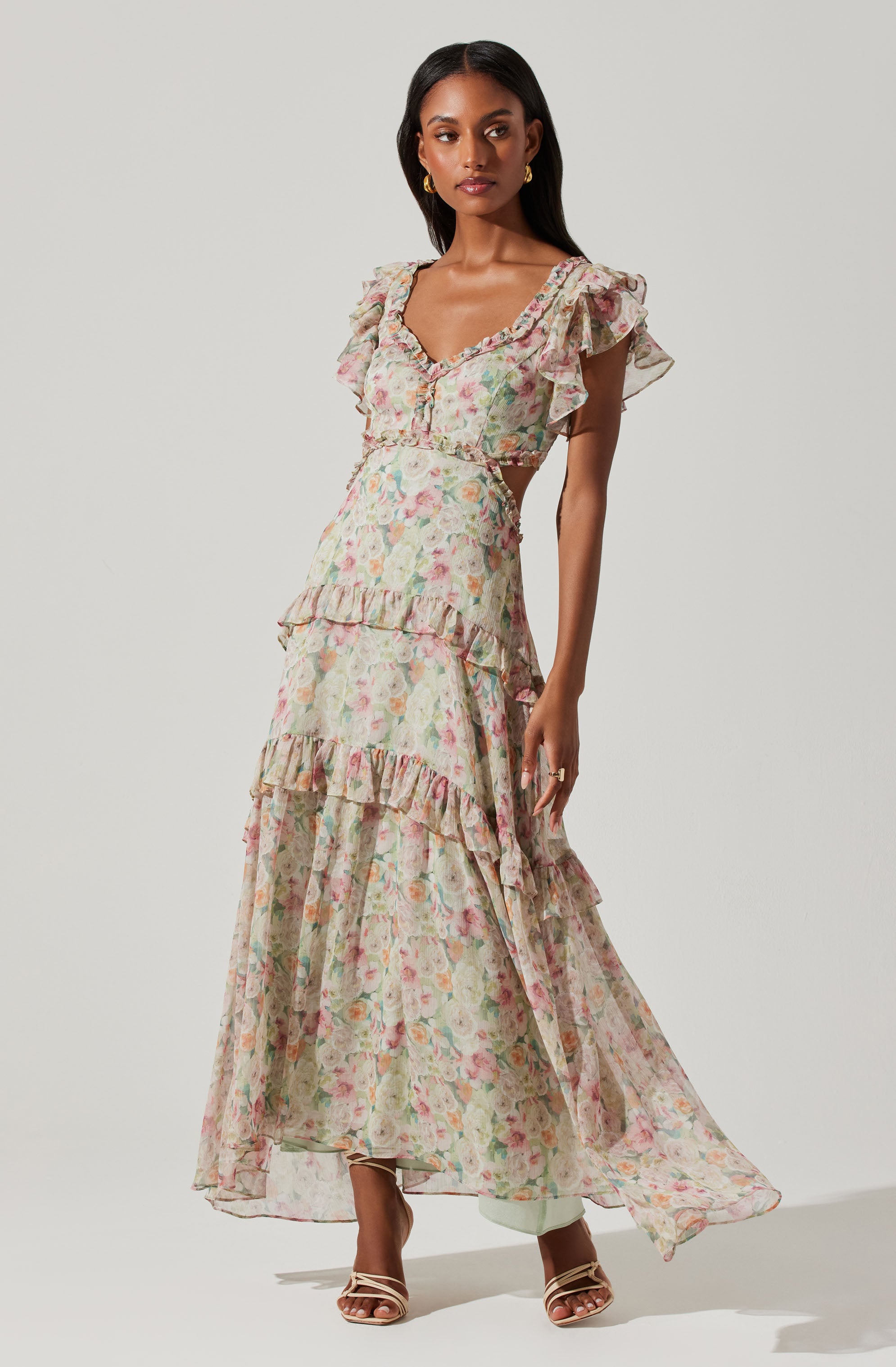 A woman is wearing a long, flowing dress with a floral pattern in soft pastel colors. The dress features ruffled details on the shoulders, along the bodice, and across the tiers of the skirt. It has short sleeves with ruffles, a V-neckline, and cutouts at the waist.