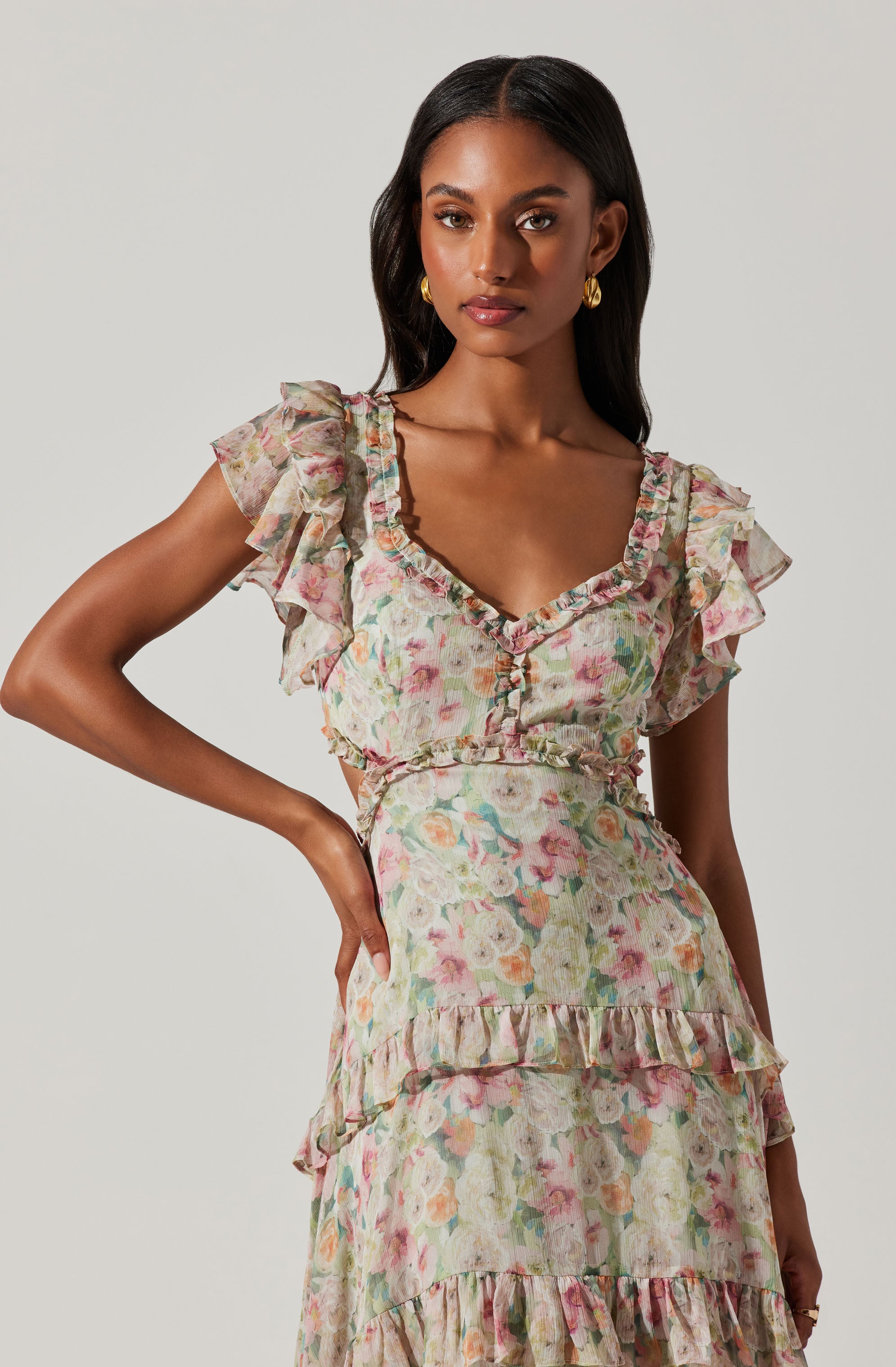 A woman is wearing a pastel-colored floral dress with a V-neckline. The dress features ruffled cap sleeves, a fitted waist, and the skirt has horizontal ruffles creating a tiered effect. The fabric has a light, airy texture with a soft floral print in shades of pink, green, and cream.