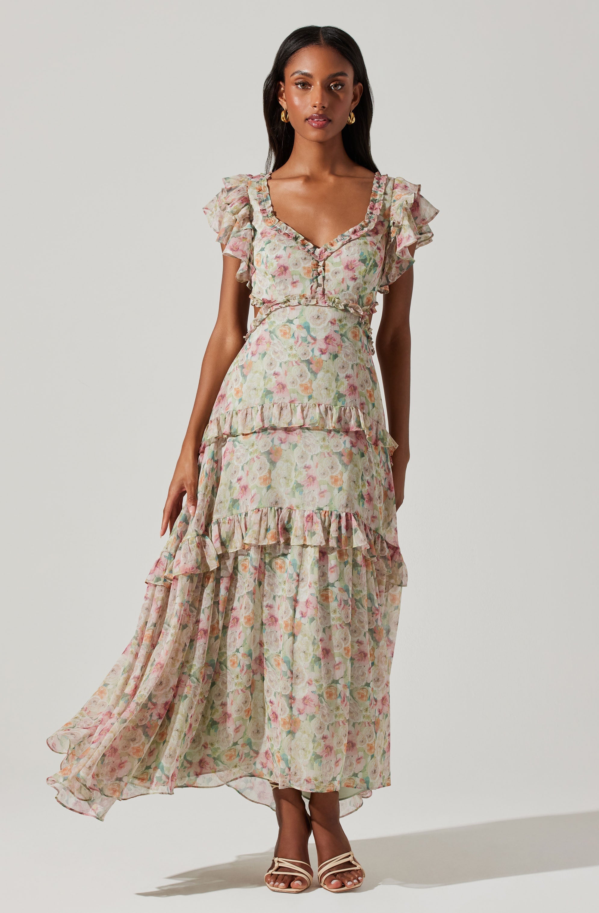 A woman is wearing a long, flowy dress with a floral pattern in pastel colors. The dress features ruffled short sleeves, a V-neckline with button details, and multiple tiered ruffles along the skirt. The fabric appears lightweight and semi-sheer. She is accessorized with gold earrings and nude strappy heels.