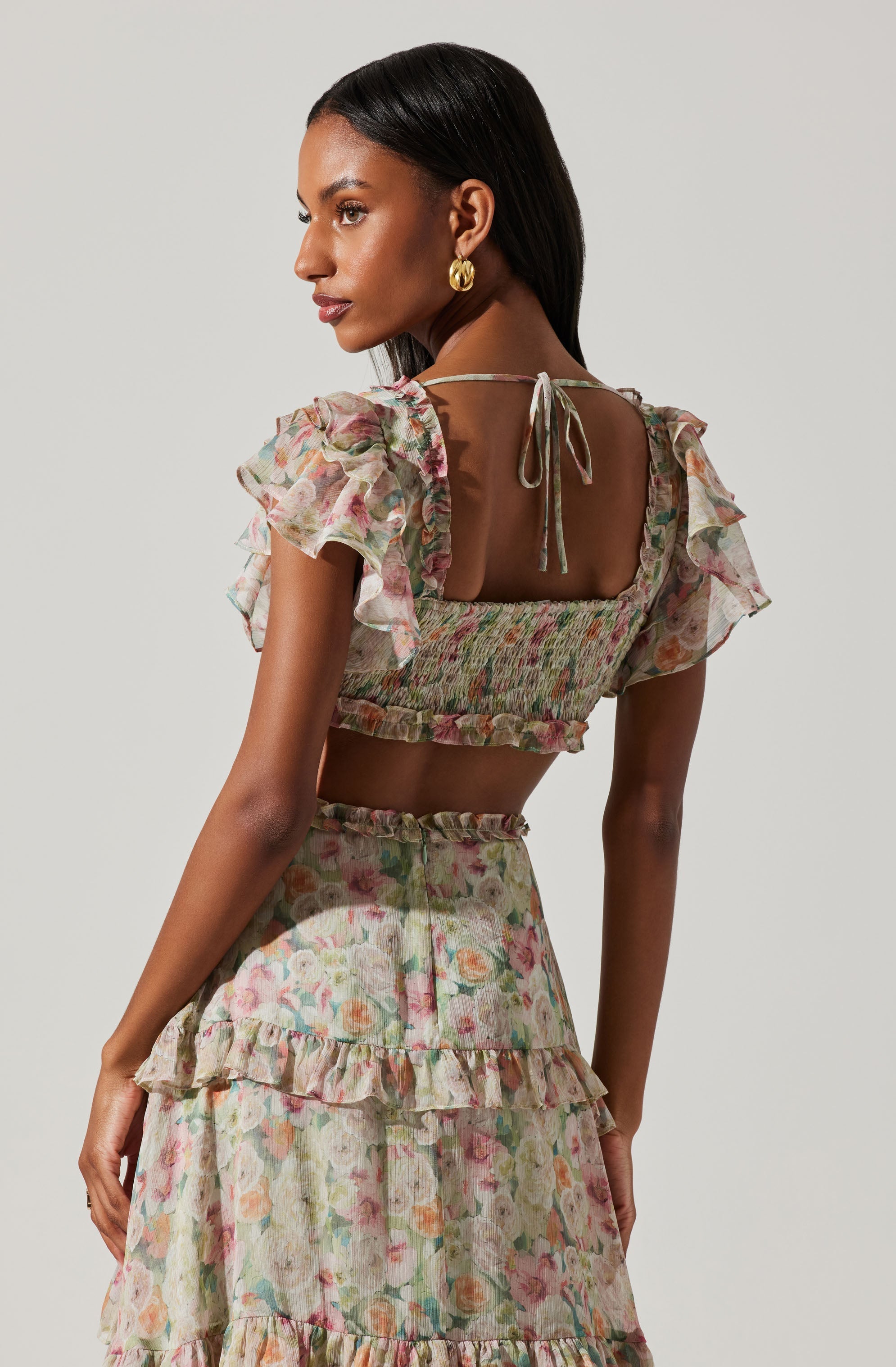 A woman is wearing a floral-print crop top with short, ruffled sleeves and a square neckline, tied at the back with a thin string. She is also wearing a matching high-waisted skirt with layers of ruffles, all in a pastel-colored floral pattern.