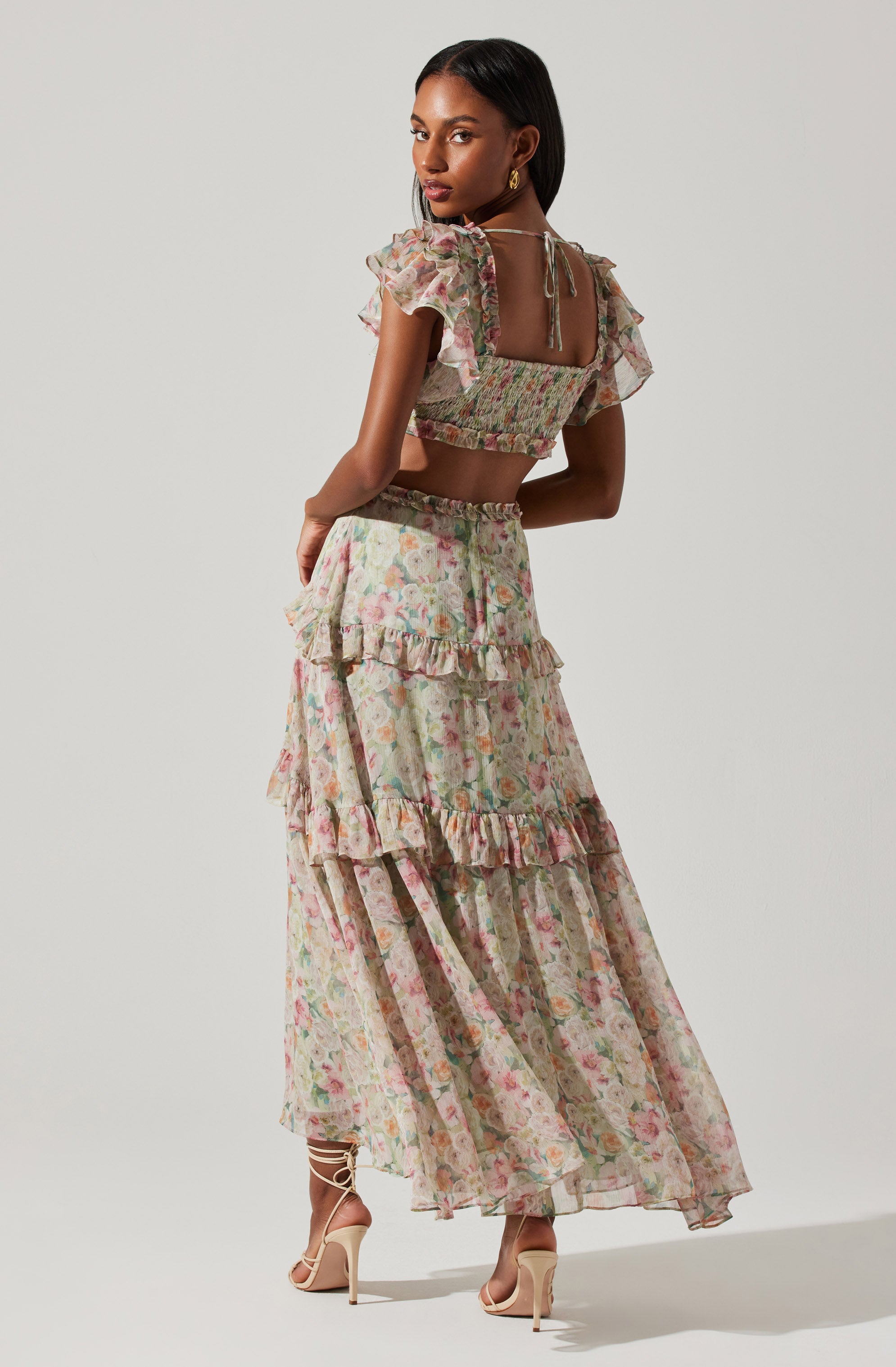A woman is wearing a two-piece floral outfit consisting of a cropped top with ruffled sleeves and an open back tied with a string, paired with a long, flowing skirt featuring multiple layers of ruffles. The fabric is light and sheer, with a pastel floral pattern. She is wearing beige high-heeled sandals with lace-up details around her ankles.