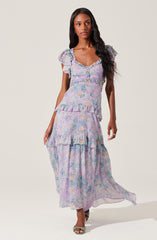 A woman is wearing a long, flowing dress with a pastel floral print in shades of purple, blue, and orange. The dress features short, ruffled sleeves, a fitted bodice with a sweetheart neckline, and ruffle details along the hem and tiers of the skirt.