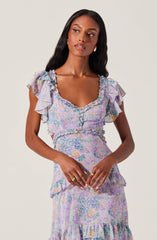 A woman is wearing a pastel-colored dress with a floral and abstract pattern. The dress features ruffled cap sleeves, a ruffled neckline, and a fitted waist. The skirt has a tiered design with ruffles at the hem.