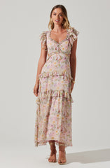 A woman is wearing a long, flowy maxi dress with a pastel floral pattern. The dress features ruffled cap sleeves, a square neckline with a small button detail, and tiered ruffles at the waist and hem. The fabric appears lightweight and breezy, suitable for warm weather. She is also wearing open-toed heels and gold jewelry.