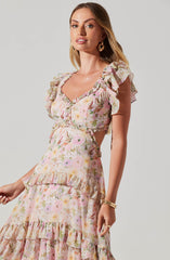 A woman wearing a pastel-colored floral dress with ruffled sleeves and tiered ruffle details. The dress features a fitted bodice with a sweetheart neckline and a flowing skirt with multiple ruffle layers. She accessorizes with gold hoop earrings and gold bangles.