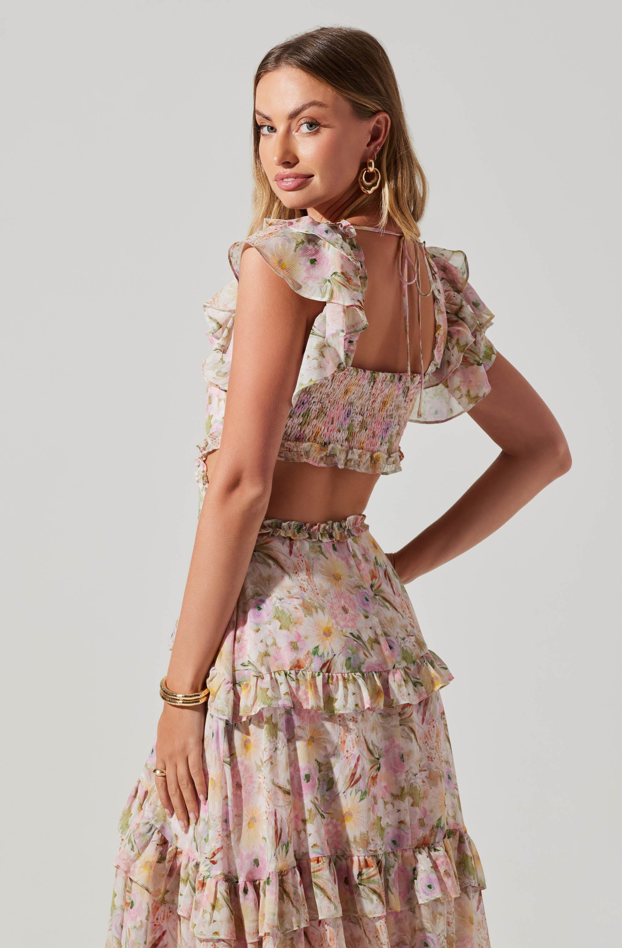 A woman is wearing a two-piece outfit with a floral pattern. The top is a sleeveless, square-neck crop top with ruffled shoulder straps and a smocked, fitted bodice with a tie at the back. The skirt is a high-waisted, ankle-length piece featuring multiple layers of ruffles, all in a pastel-colored floral print.