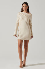 A woman is wearing a cream-colored, long-sleeved mini dress with a subtle textured pattern. The dress features a relaxed fit with a draped neckline that extends over one shoulder. She is also wearing gold hoop earrings and beige strappy high-heeled sandals.