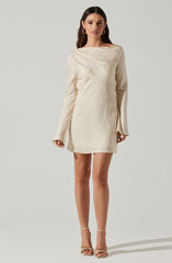 A woman is wearing a cream-colored, long-sleeved dress with a subtle textured pattern. The dress has a loose, relaxed fit with a boat neckline and slightly flared cuffs on the sleeves. She is accessorizing with large hoop earrings and high-heeled sandals with ankle straps.