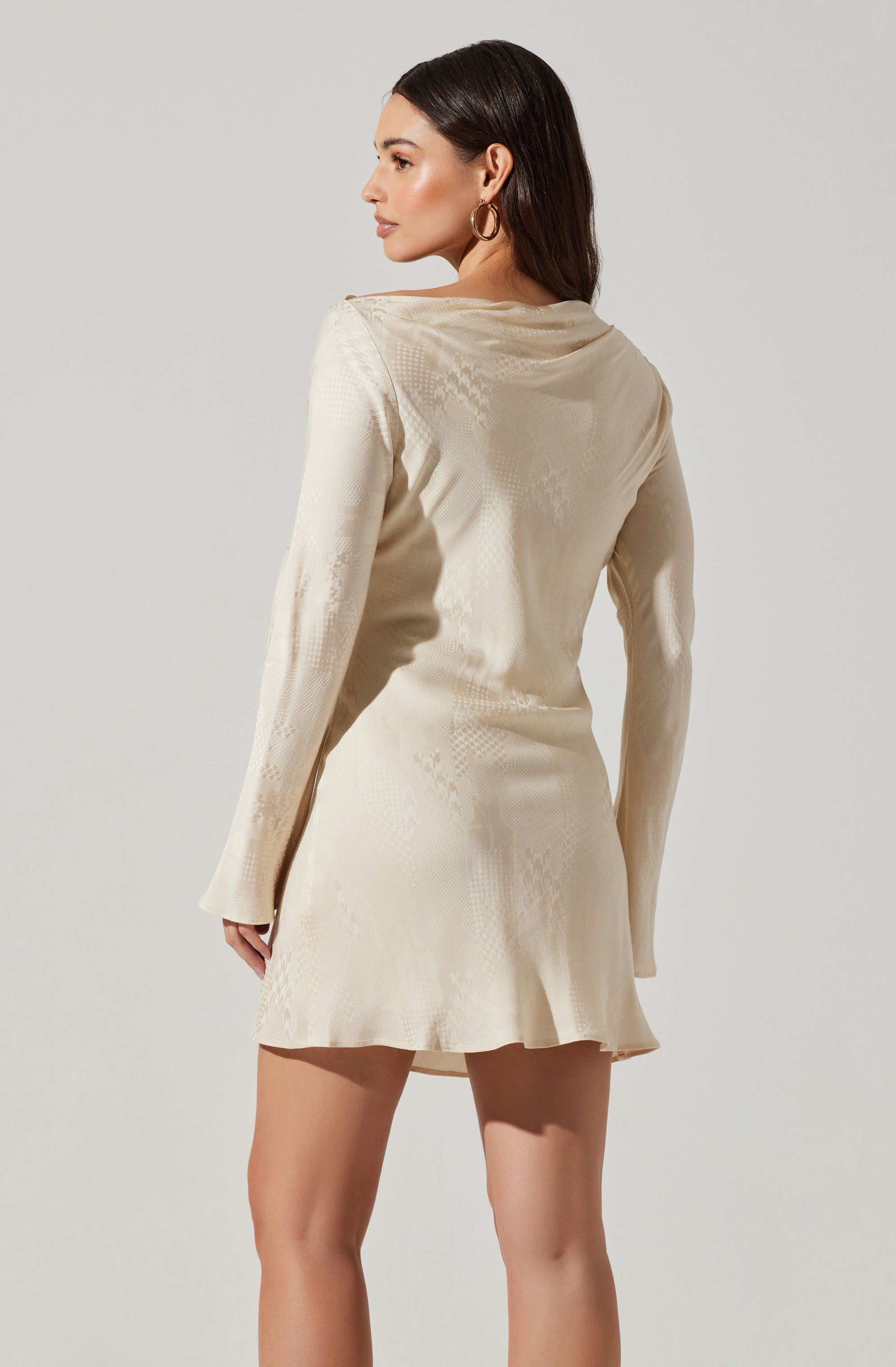 A woman is wearing a long-sleeved, cream-colored dress with a subtle textured pattern. The dress has a loose, slightly draped neckline and a fitted waist, flaring out into a short, flowy skirt.