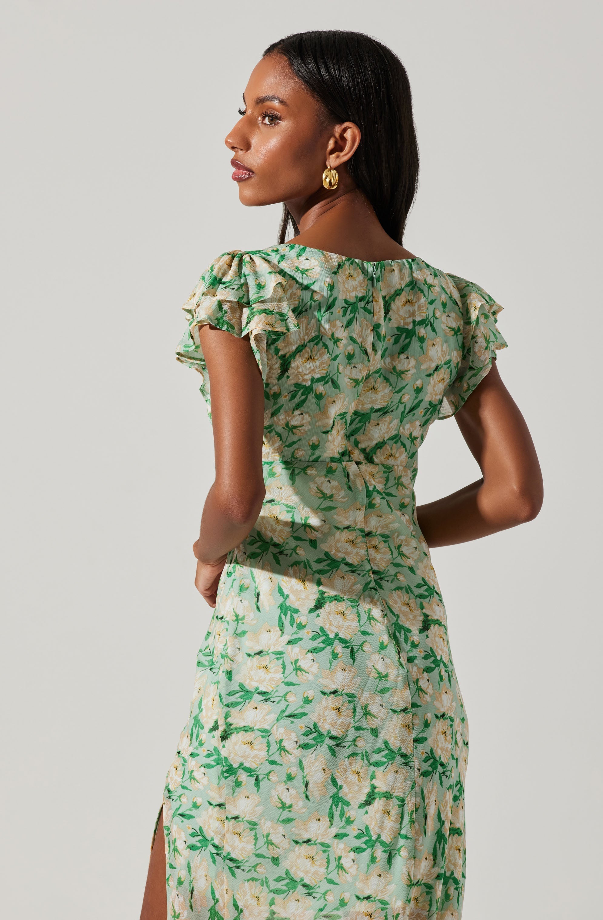 A woman is wearing a light-colored, floral dress with green leaves and large, soft yellow flowers. The dress features short, ruffled sleeves and a slit on the side. The fabric appears lightweight and slightly shiny.
