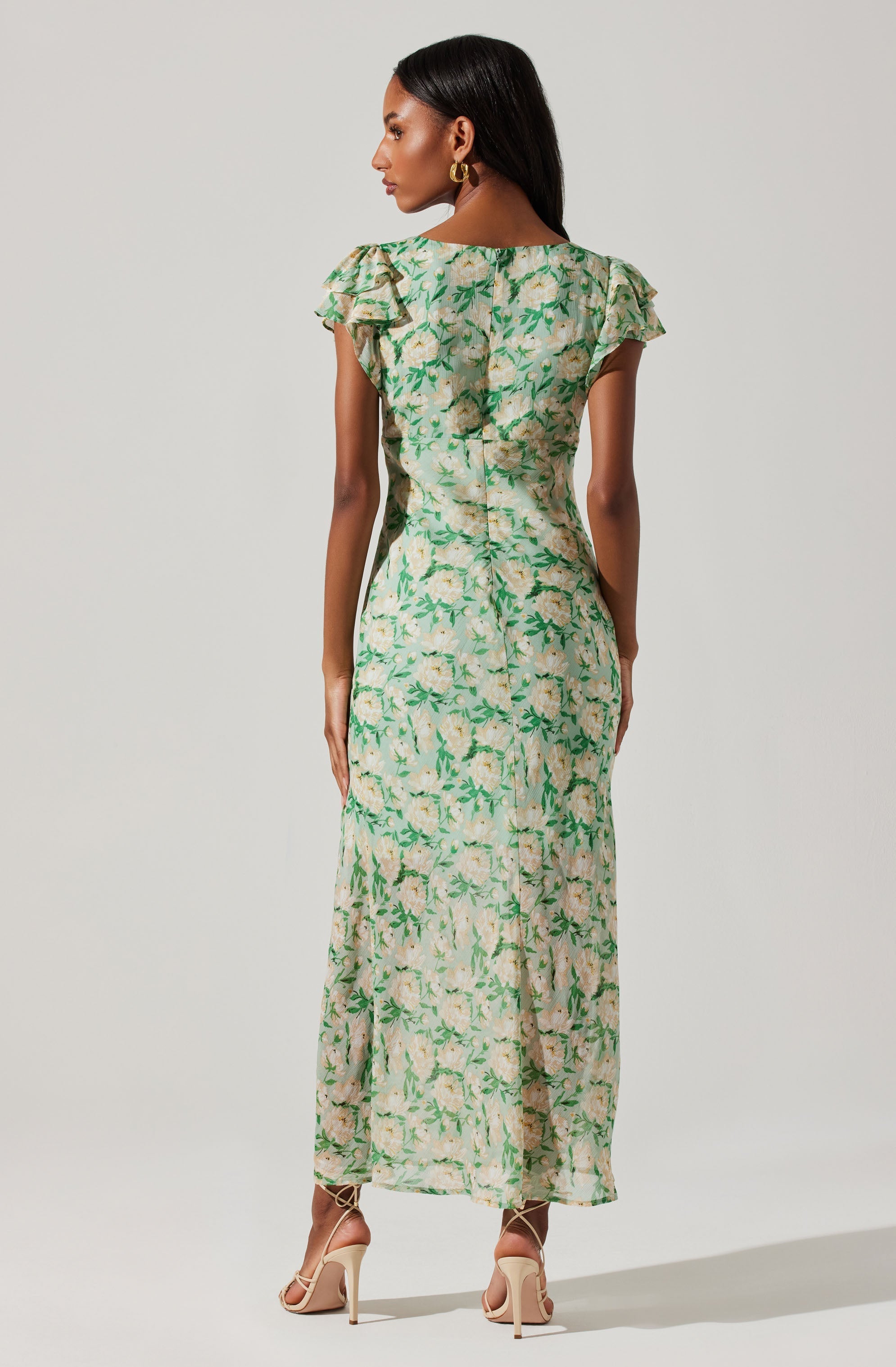A woman is wearing a long, flowy dress with a floral pattern. The dress has a light green and beige background with darker green leaves and light-colored flowers. It features short, ruffled sleeves and a fitted waist, extending down to her ankles. She is also wearing nude high-heeled sandals.