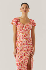 A woman is wearing a pink floral dress with short, ruffled sleeves. The dress has a fitted bodice with a sweetheart neckline and a front slit that reveals her leg. The floral pattern features shades of pink, orange, and yellow.