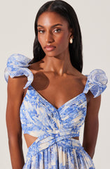 A woman is wearing a light blue and white patterned dress with ruffled shoulder straps, a sweetheart neckline, and cutouts at the waist. The dress has a pleated bodice and a flowing skirt with a matching pattern.