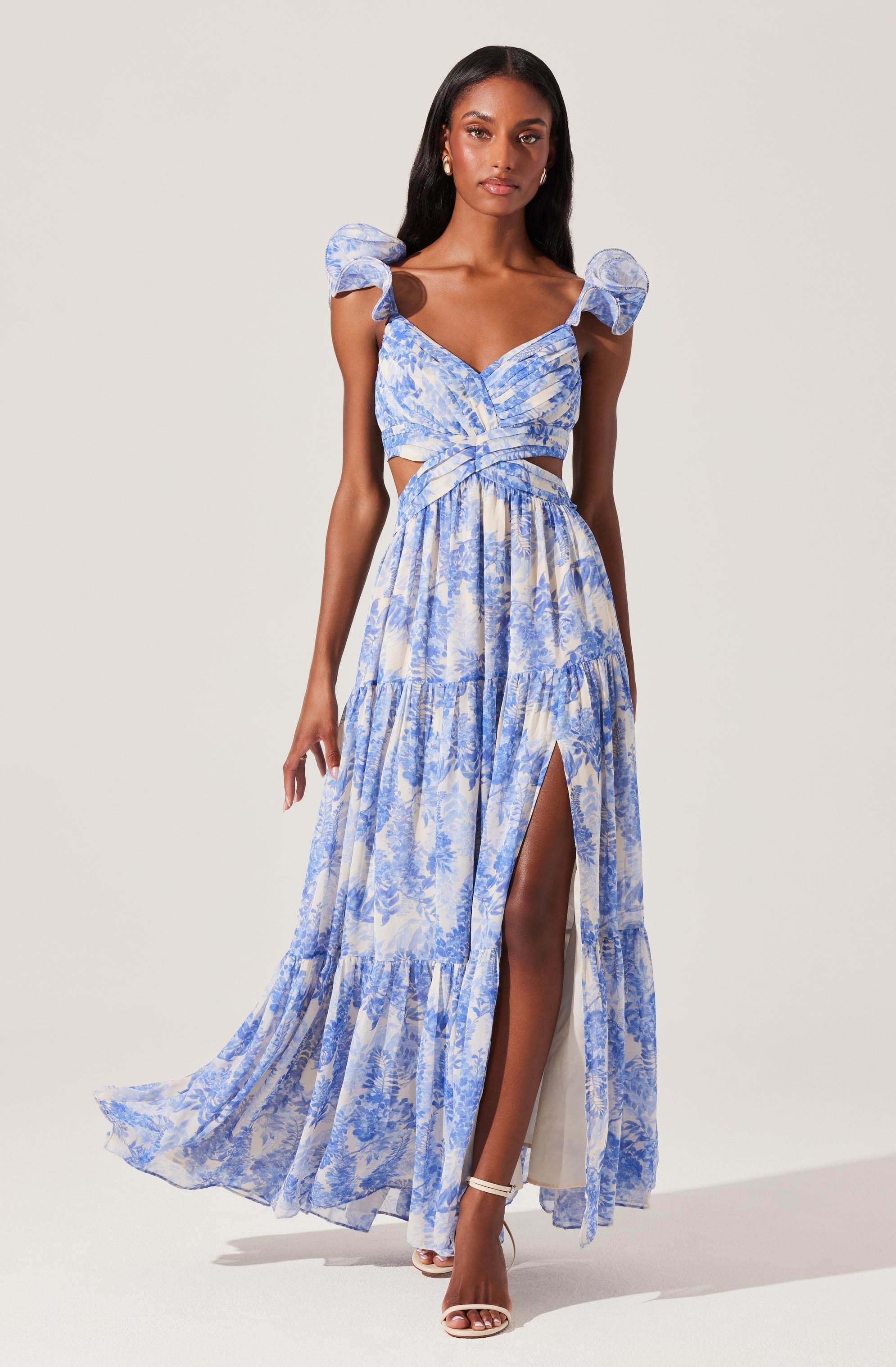 A woman is wearing a light blue and white floral maxi dress with a fitted bodice and puffed short sleeves. The dress features a sweetheart neckline, cut-out sides at the waist, and tiered skirt with a high slit. She is also wearing high-heeled sandals.
