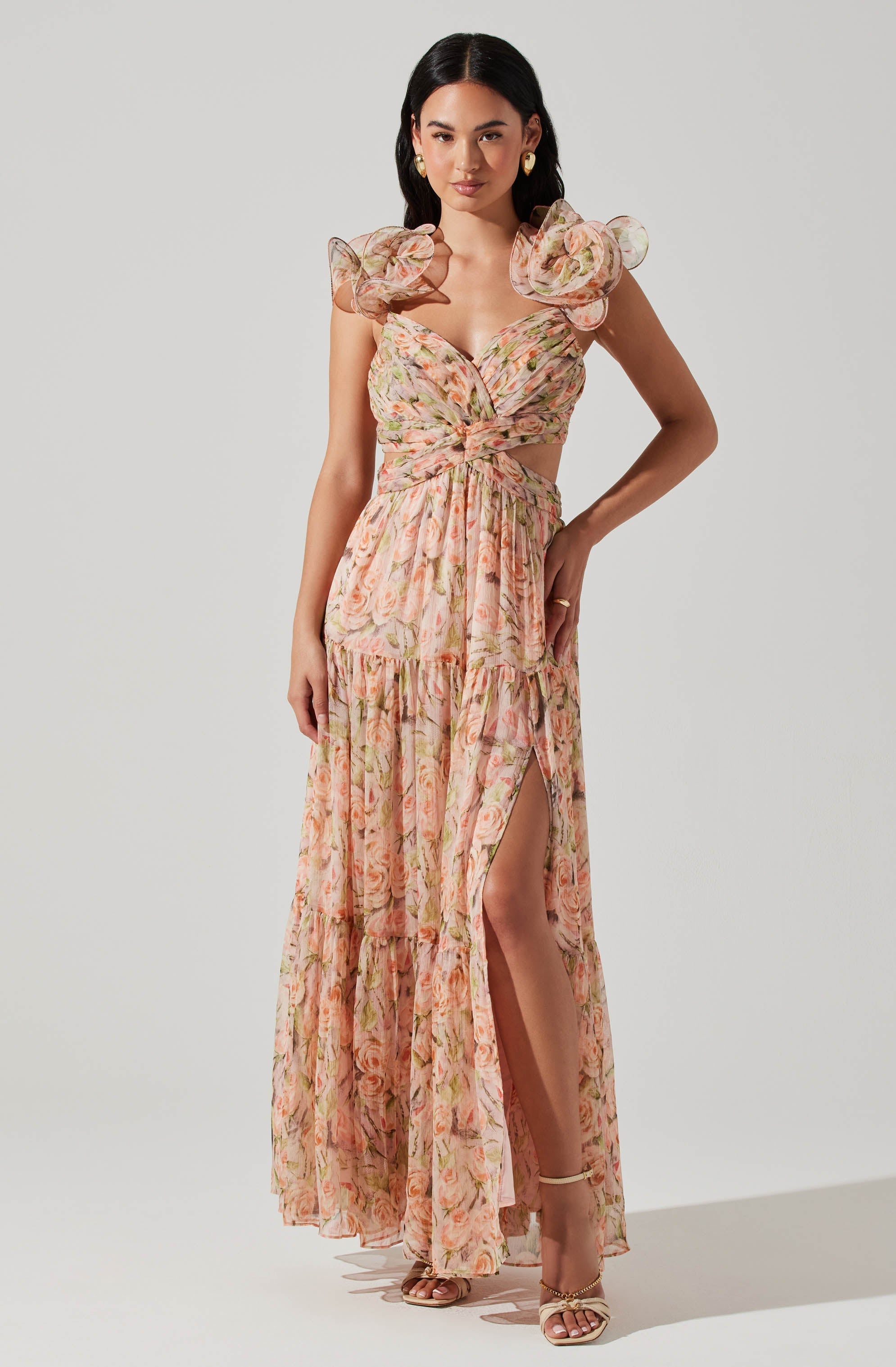 A woman is wearing a floral dress with a light, pastel-colored pattern. The dress features ruffled, puffed sleeves and a bustier-style top with a knot detail at the waist. It has a high slit on one side, revealing her leg and gold strappy heels.