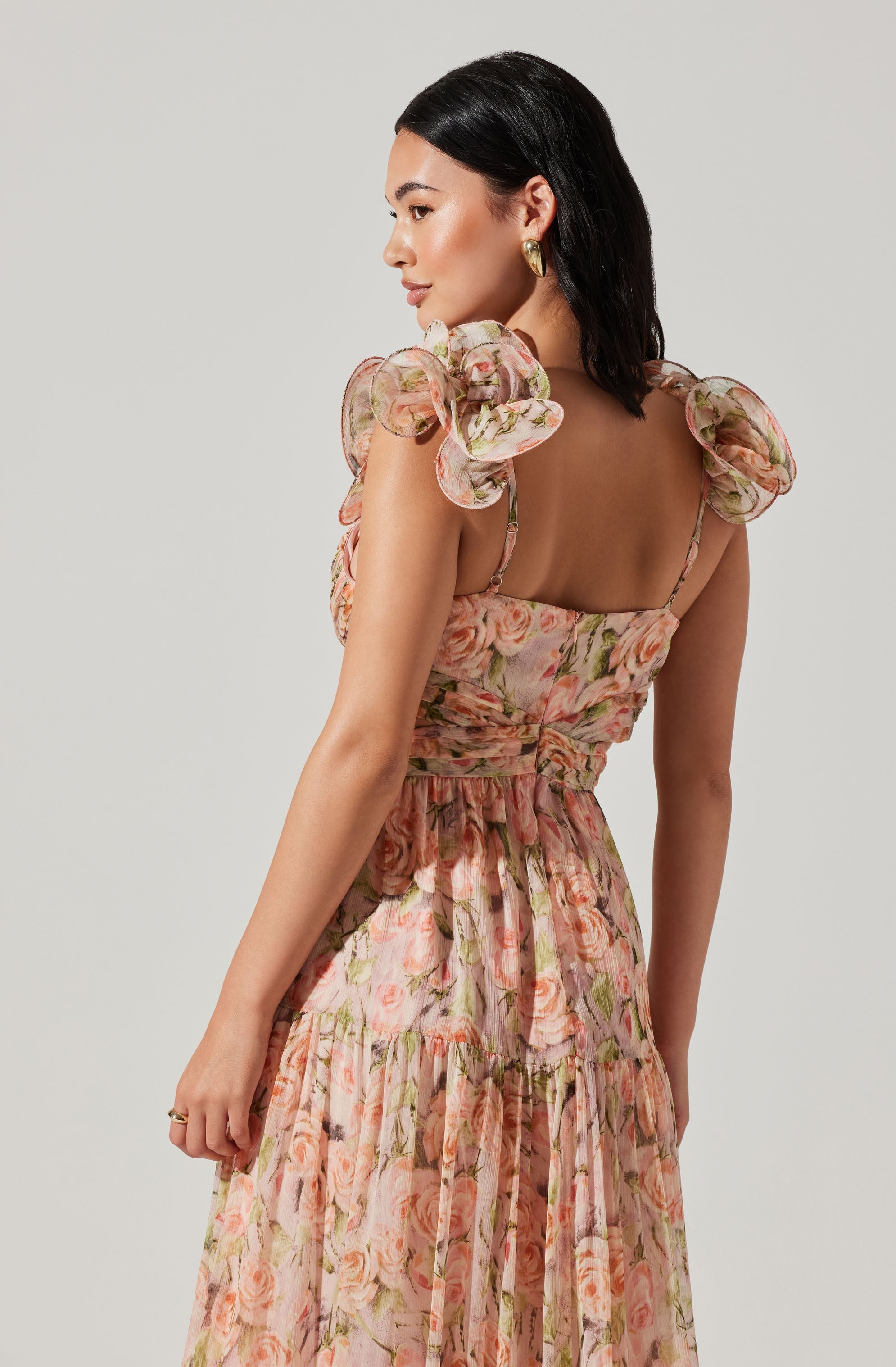 A woman is wearing a sleeveless, floral dress with a fitted bodice and a full, tiered skirt. The dress features shoulder details with large, ruffled, puffed sleeves made from a sheer, lightweight fabric with a floral pattern. The fabric of the dress has a pink and peach floral print with green leaves.