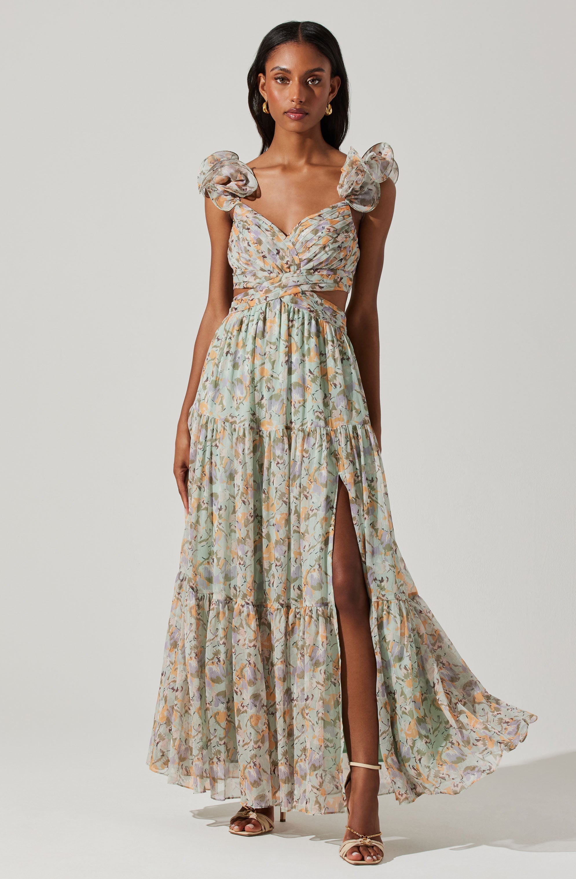 A woman is wearing a long, flowy dress with a floral pattern in soft pastel shades. The dress features puffed, ruffled sleeves, a fitted bodice with a twist detail at the front, and a high slit on one side, revealing her leg. She is also wearing strappy heels.