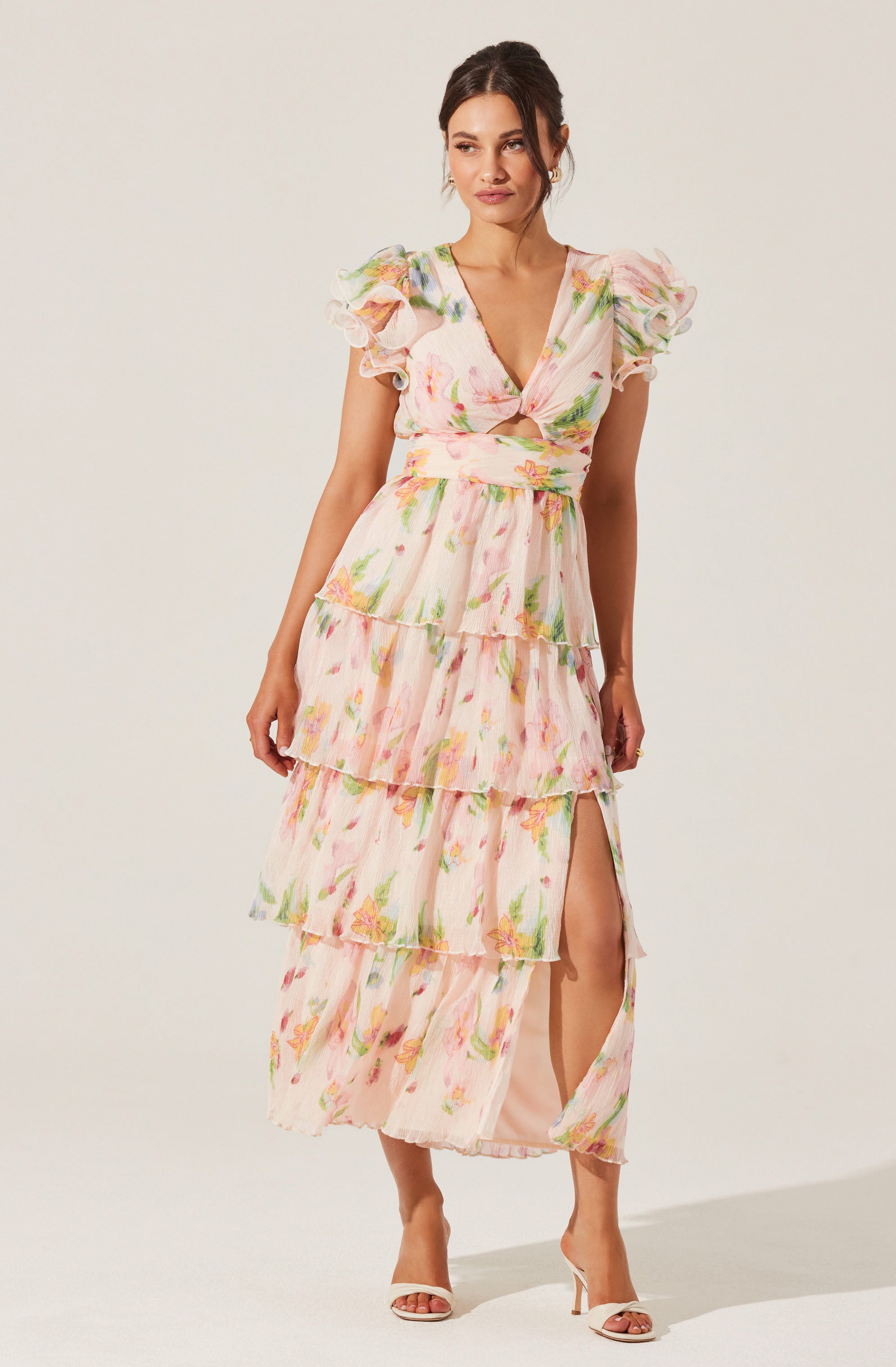 A woman is wearing a long, flowing dress with a pastel floral pattern. The dress features short, puffed sleeves with ruffled edges, a V-neckline, and a fitted waist. It has multiple layered ruffles descending to the hem, with a slit on the left side revealing her leg. She is also wearing white high-heeled sandals.