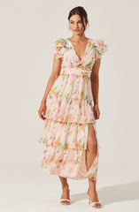A woman is wearing a long, flowing dress with a pastel floral pattern. The dress features short, puffed sleeves with ruffled edges, a V-neckline, and a fitted waist. It has multiple layered ruffles descending to the hem, with a slit on the left side revealing her leg. She is also wearing white high-heeled sandals.