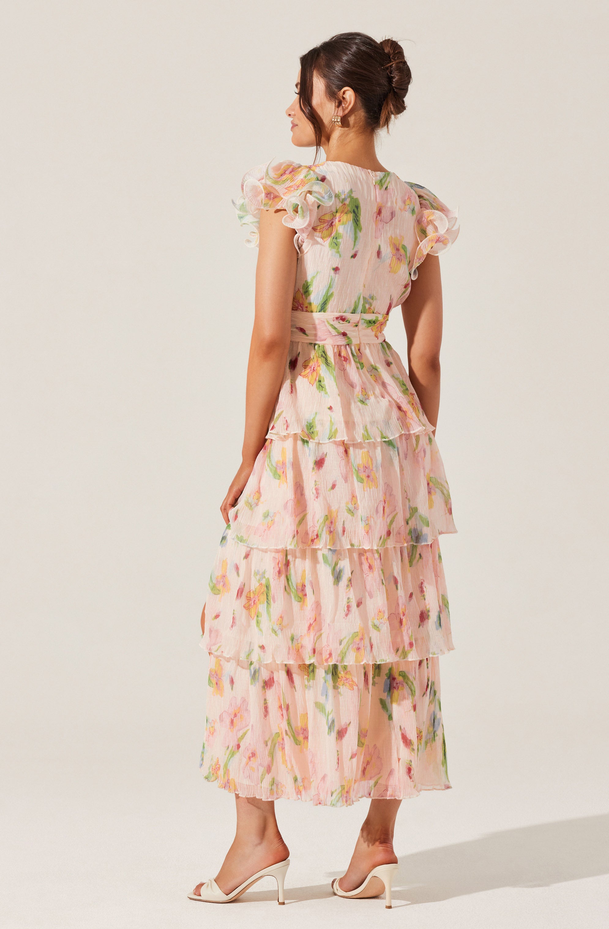 A woman is wearing a long, flowy dress with a pastel floral pattern. The dress features a high waistline with a matching fabric belt, and the fabric is lightweight and semi-sheer. The dress has short, ruffled sleeves with layered, tiered skirts that create a tiered effect from the waist down. She is also wearing high-heeled shoes in a neutral color.