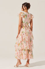 A woman is wearing a long, flowy dress with a pastel floral pattern. The dress features a high waistline with a matching fabric belt, and the fabric is lightweight and semi-sheer. The dress has short, ruffled sleeves with layered, tiered skirts that create a tiered effect from the waist down. She is also wearing high-heeled shoes in a neutral color.