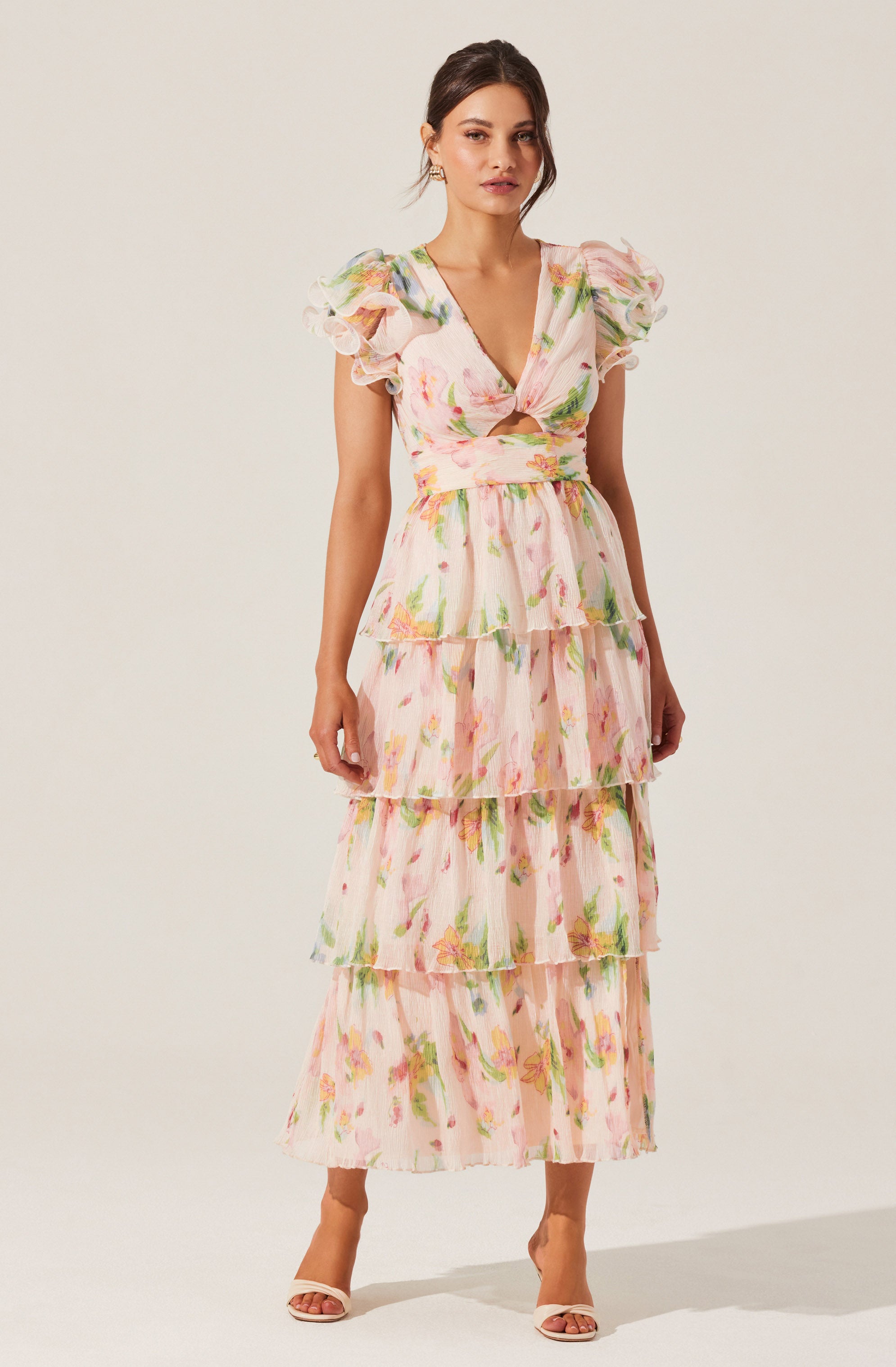 A woman is wearing a long, tiered dress with a floral pattern featuring pink, yellow, and green colors. The dress has a deep V-neckline with a twisted knot detail at the bust, short puff sleeves.