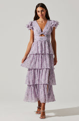 A woman is wearing a light purple, floral-patterned dress with multiple layers of ruffles. The dress has a deep V-neckline with a twist knot detail at the front, creating a small cutout at the waist. The short, puffed sleeves are made of ruffled fabric, adding volume. She is also wearing open-toe heels with ankle straps.