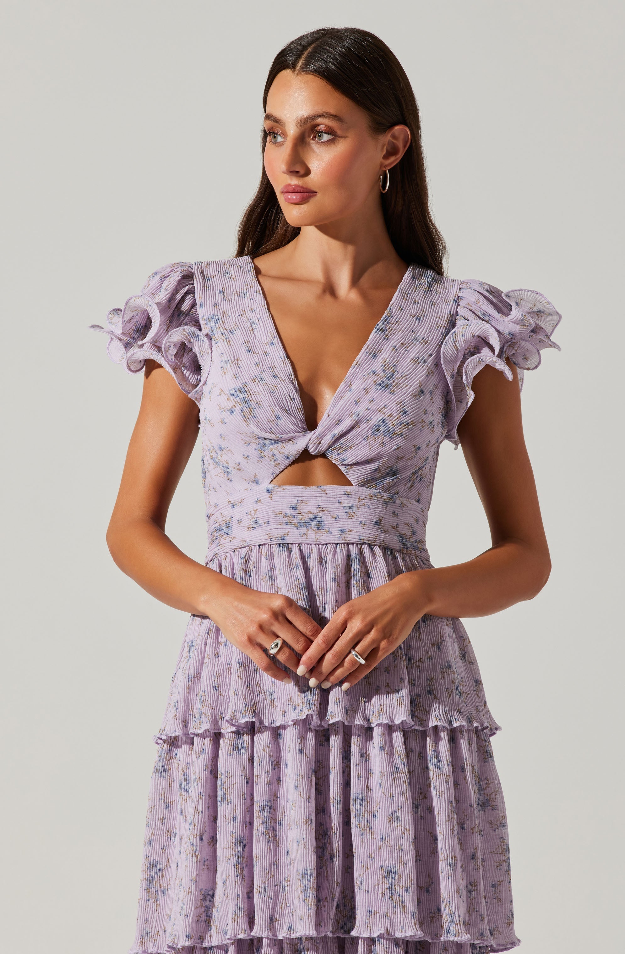 A woman is wearing a light purple dress with a floral pattern. The dress features puffed, ruffled short sleeves, a deep V neckline with a twist knot at the bust, and a cutout detail just below the knot. The bodice and skirt have a layered, tiered design with ruffles and vertical pleats, creating a textured, flowing appearance.