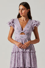 A woman is wearing a light purple dress with a floral pattern. The dress features puffed, ruffled short sleeves, a deep V neckline with a twist knot at the bust, and a cutout detail just below the knot. The bodice and skirt have a layered, tiered design with ruffles and vertical pleats, creating a textured, flowing appearance.