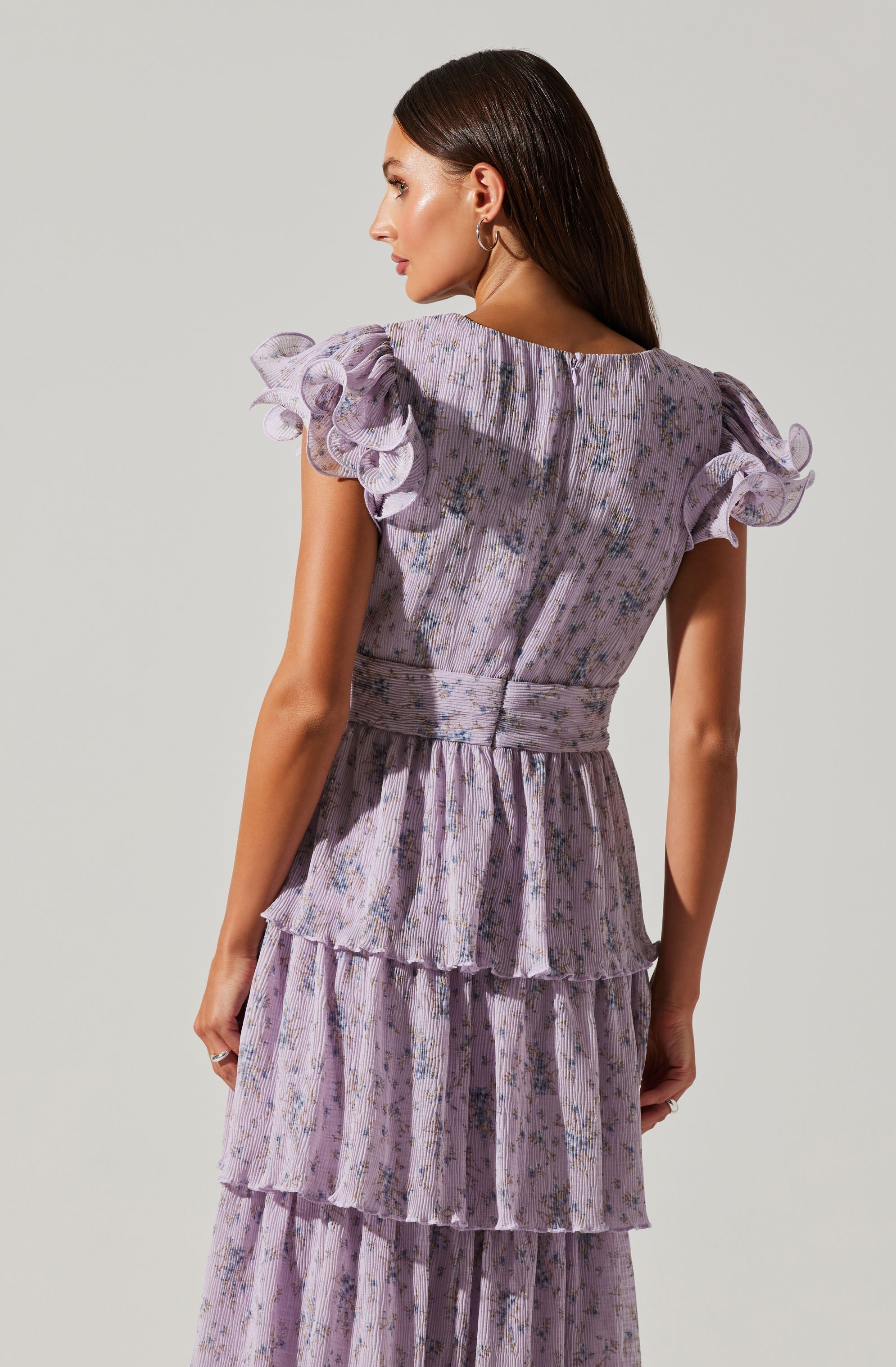 A woman is wearing a light purple dress with a floral pattern. The dress features ruffled, gathered cap sleeves with multiple layers. The fabric is textured with a subtle vertical stripe pattern, and the waist is cinched, creating a fitted silhouette. The skirt has tiered ruffles that flow down to the hem.