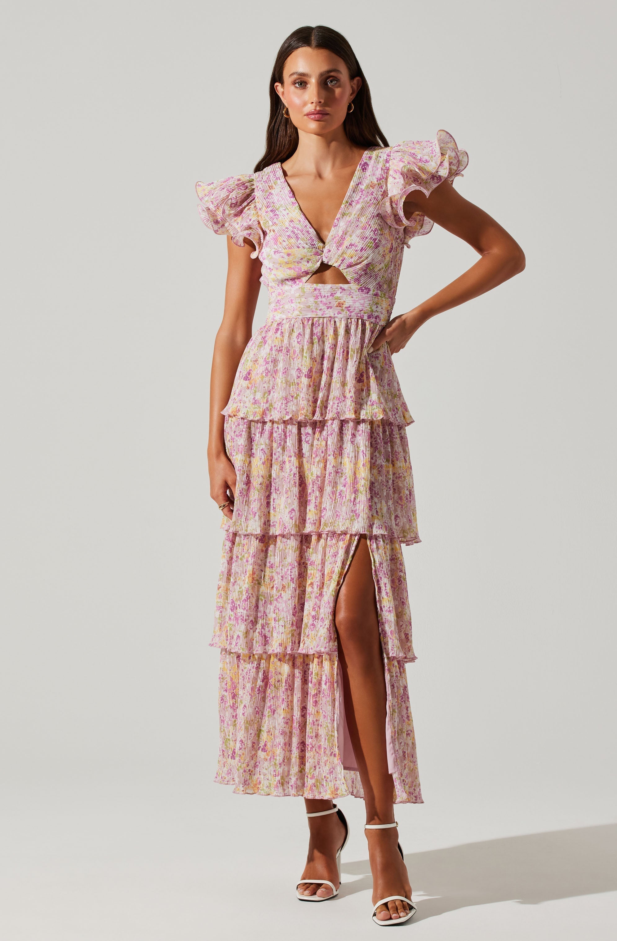 A woman is wearing a long, tiered dress with a floral pattern in pastel pink, yellow, and purple. The dress has a V-neckline with a small cutout just below the bust, puffed short sleeves with ruffled edges, and a high slit on the left side revealing her leg. She is also wearing white strappy high-heeled sandals.