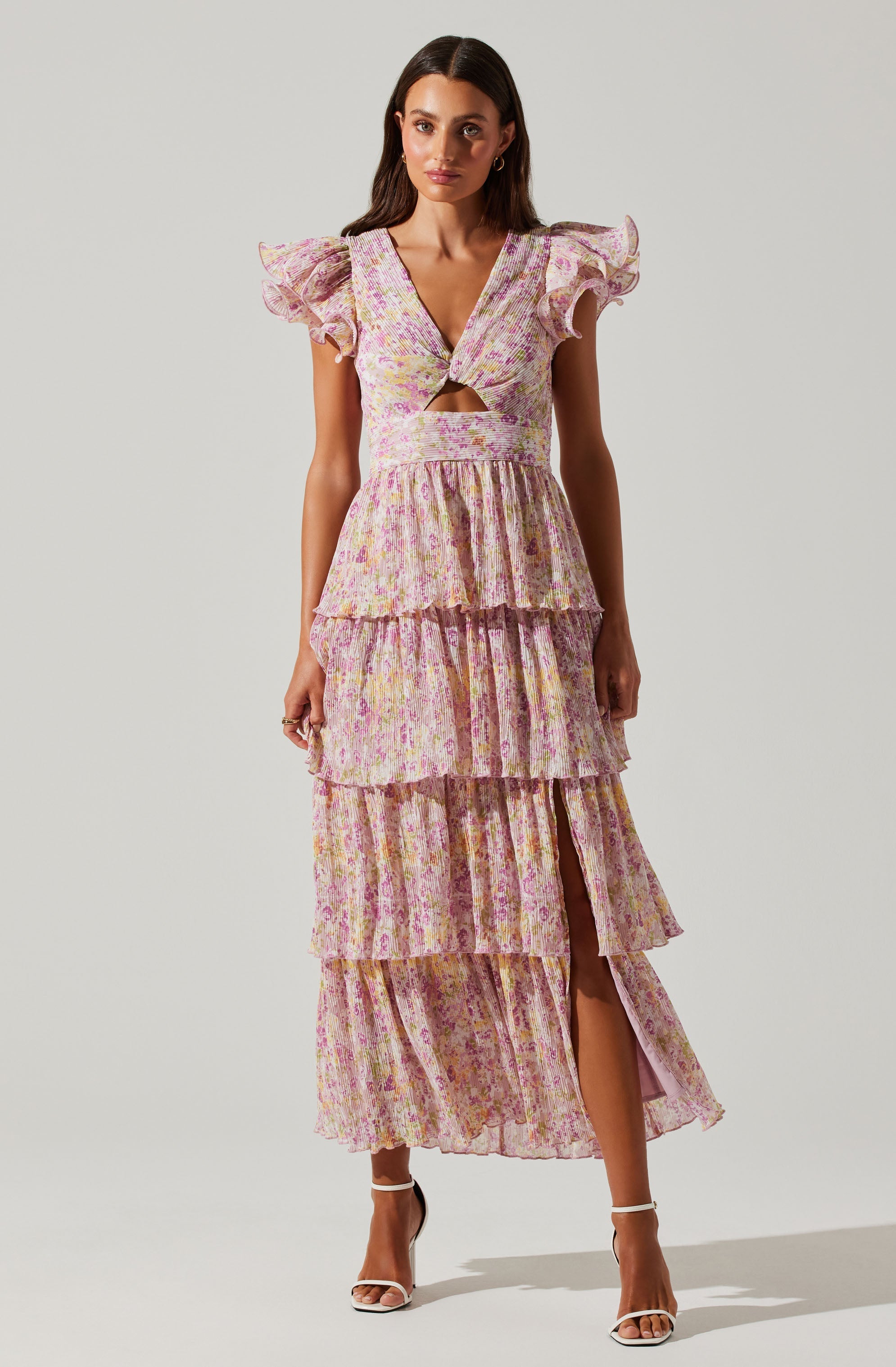 The dress is a long, flowy garment made of lightweight fabric with a pastel floral pattern in shades of pink, purple, and yellow. It features a deep V-neckline with a small cutout at the center, and has short, ruffled sleeves. The waist is fitted, and the skirt has multiple layers of tiered ruffles that extend to the ankles, with a slit on one side revealing the leg. The dress exhibits a soft, feminine, and summery style.