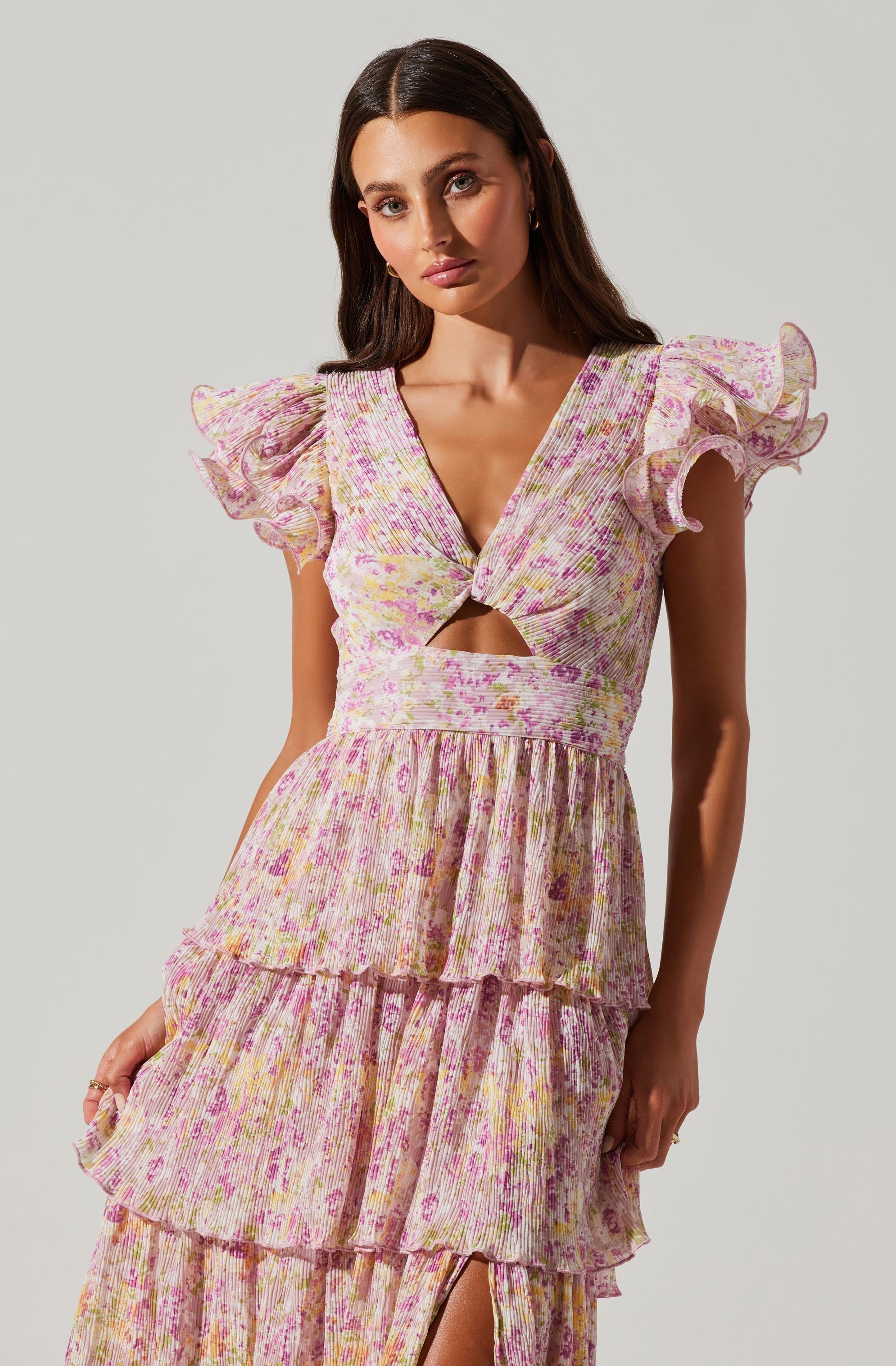 A woman is wearing a pastel-colored dress with a floral pattern. The dress features a V-neckline with a twist detail at the center, a cutout just below the bust, and ruffled, short, puffed sleeves. The skirt has multiple layers of ruffles, creating a tiered look with a slit up the front.