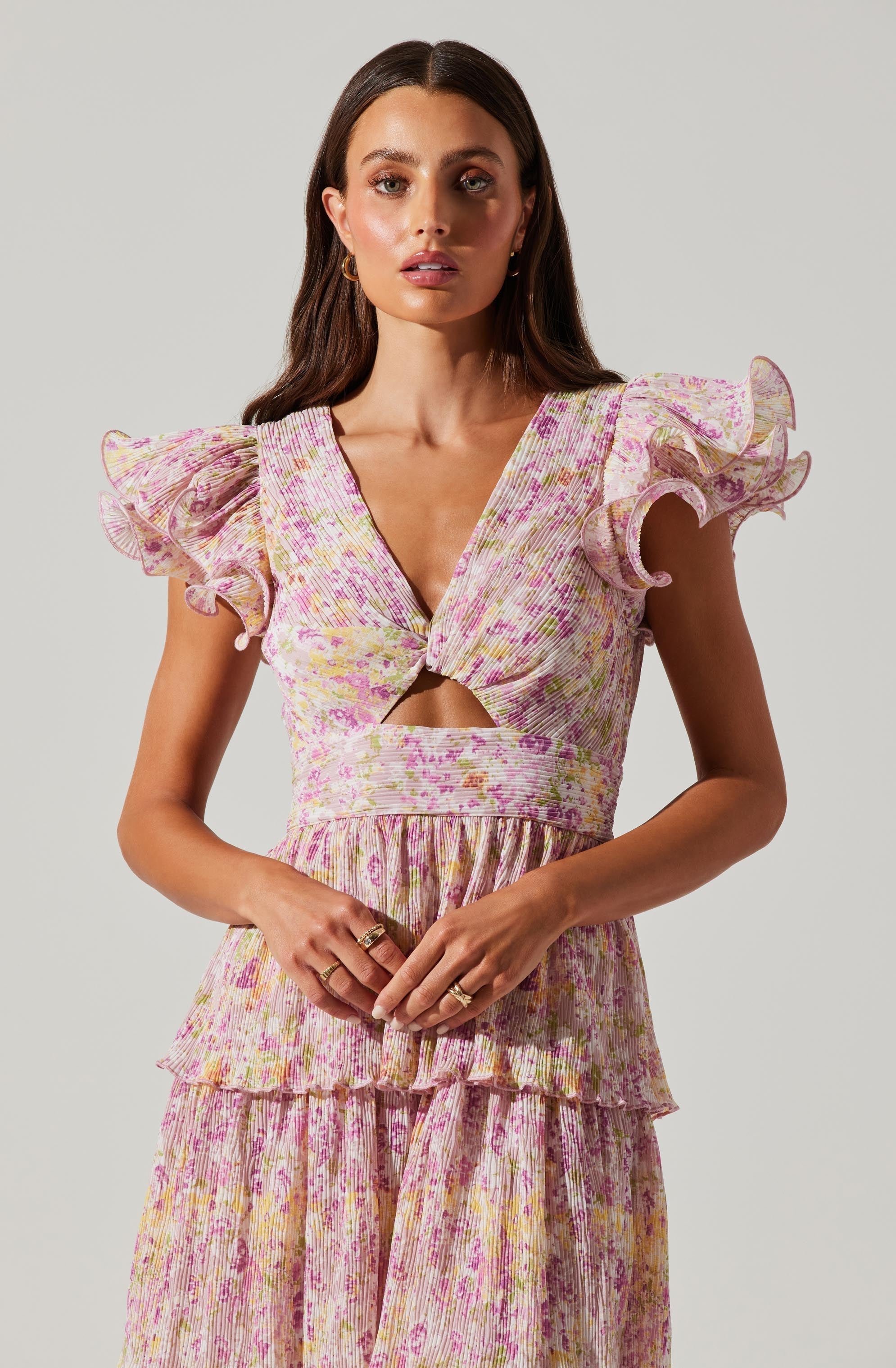 A woman is wearing a pink and yellow floral dress with a textured fabric. The dress features ruffled, puffed sleeves, a deep V-neckline, a small cutout below the bust, and a gathered, ruffled hem.