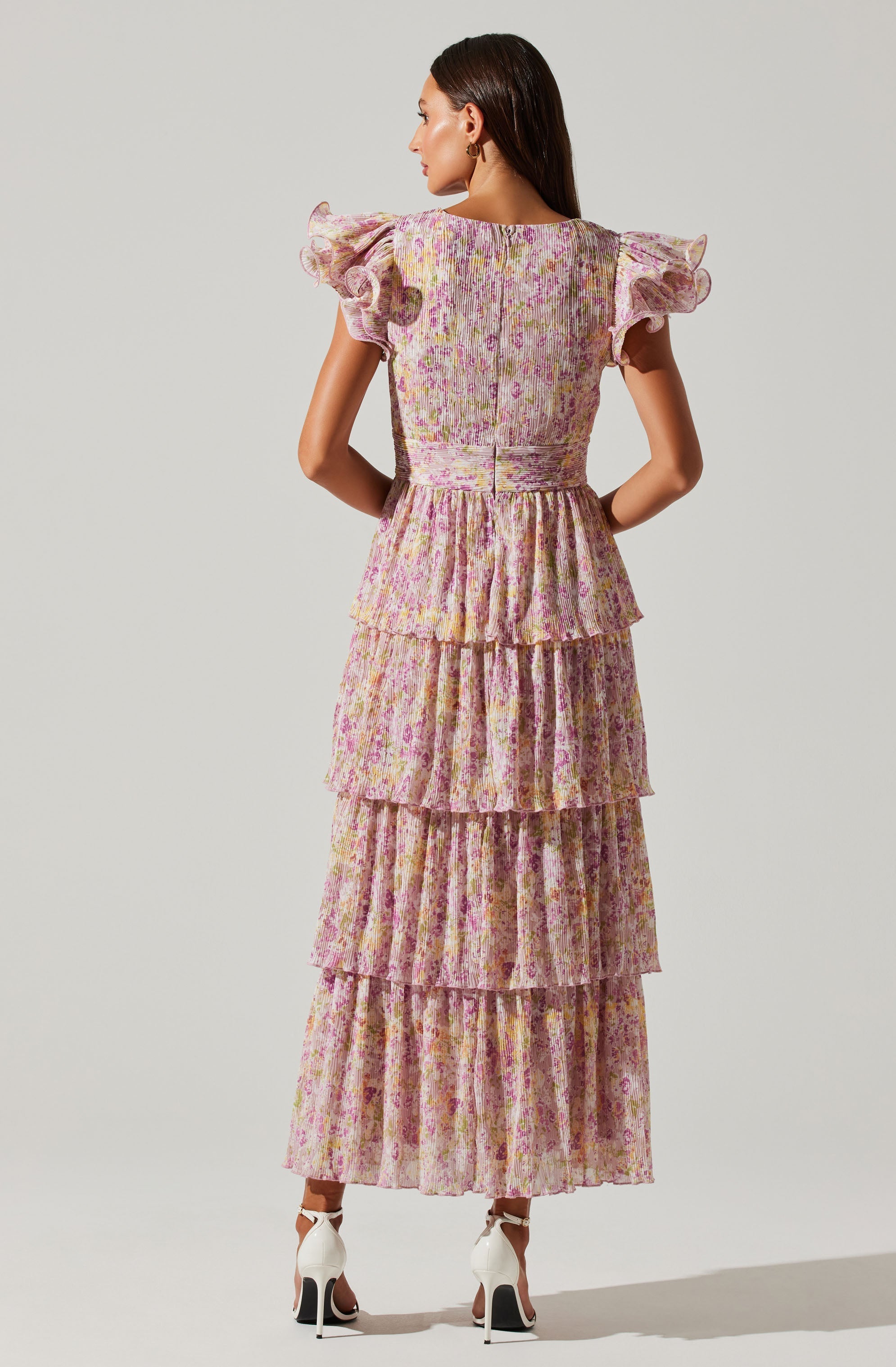 A woman is wearing a long, layered dress with a floral print. The dress features short, ruffled sleeves and a fitted waist, with multiple tiered ruffles making up the skirt. The fabric appears lightweight and slightly textured, with a pattern of pink and purple flowers on a muted background. She is also wearing white high-heeled shoes.
