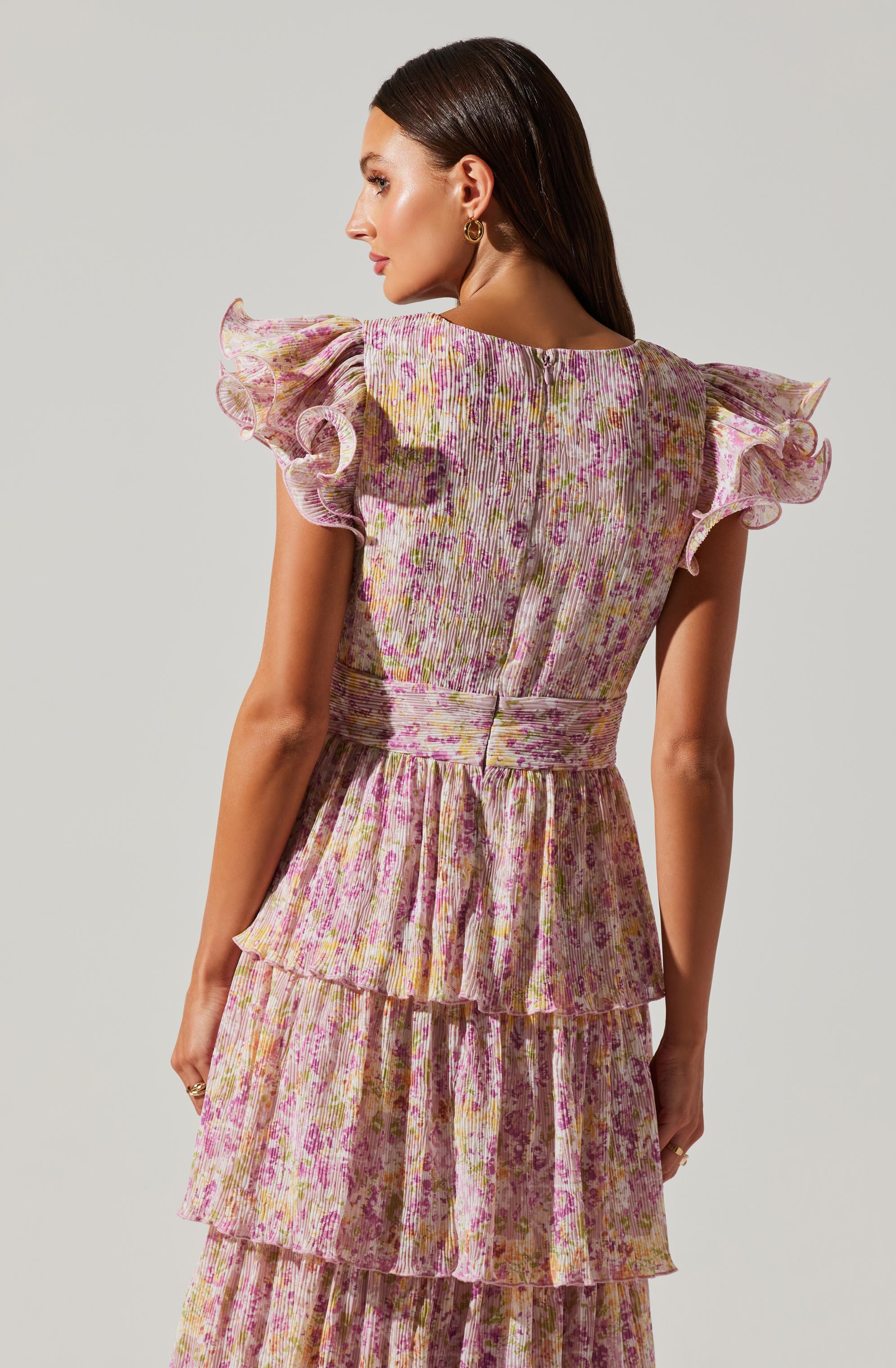 A woman is wearing a dress with ruffled short sleeves, a fitted waist, and a tiered skirt. The fabric has a pleated texture with a floral pattern in shades of pink, purple, and yellow.