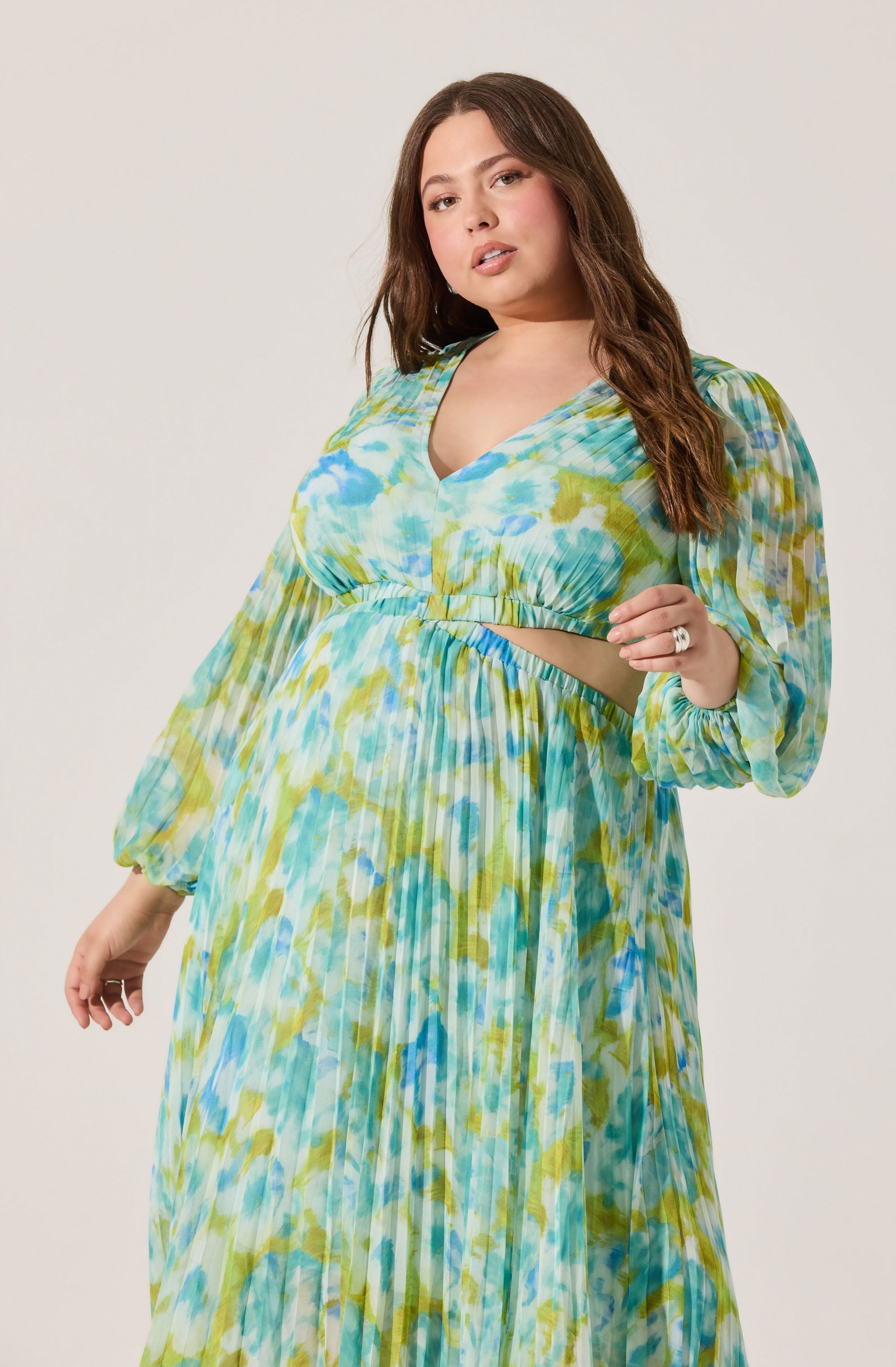 A woman is wearing a long, flowing dress with a multicolored, watercolor-style pattern in shades of blue, green, and yellow. The dress has long, puffed sleeves with elastic cuffs, a V-neckline, and a cutout at the midriff.