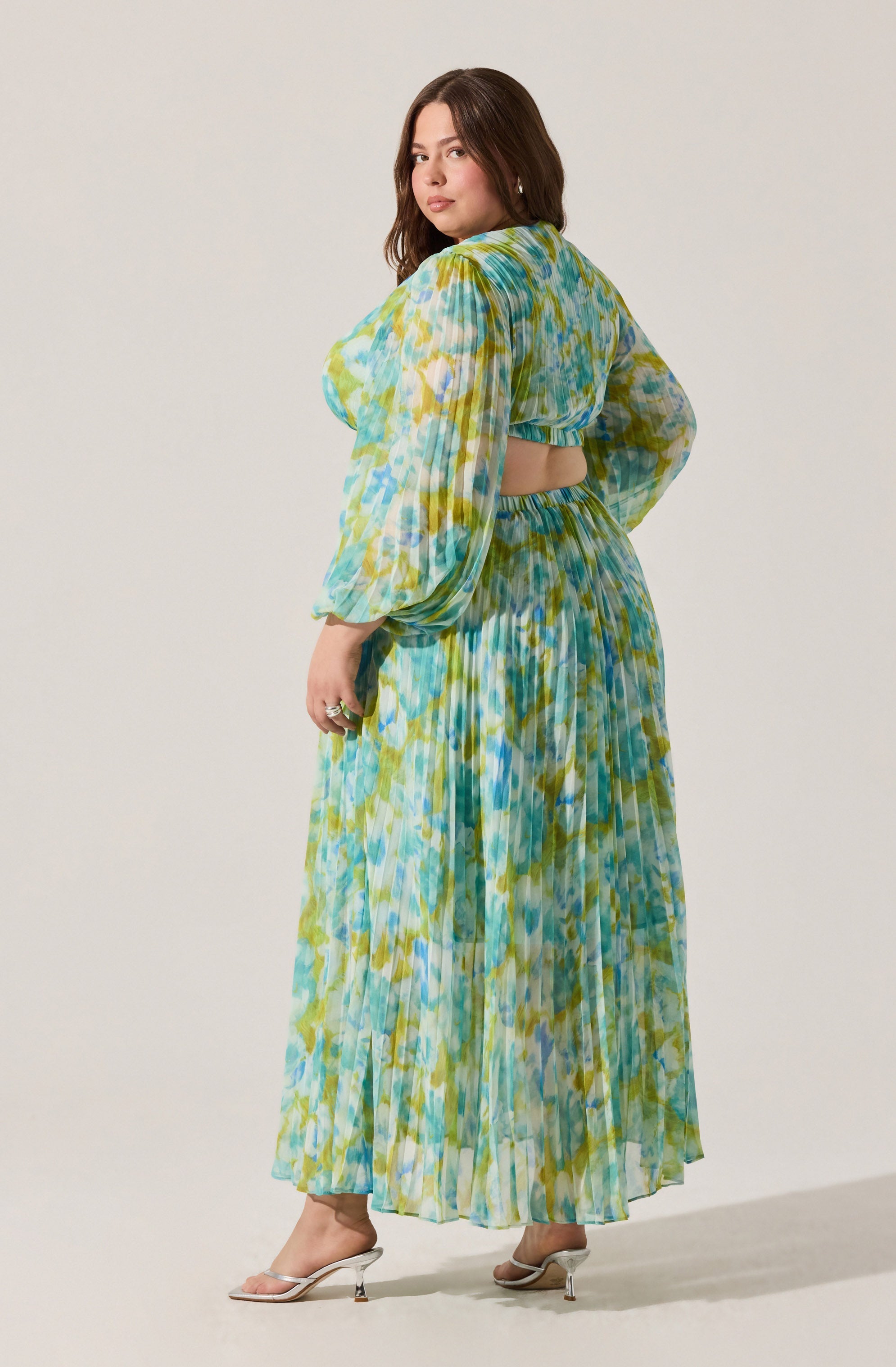 A woman is wearing a flowy, two-piece outfit with a multicolored abstract pattern in shades of blue, green, and yellow. The top has long, puffed sleeves and a cut-out detail at the back, revealing her midriff. The skirt is high-waisted, pleated, and reaches down to her ankles. She is also wearing light-colored, open-toe heels.