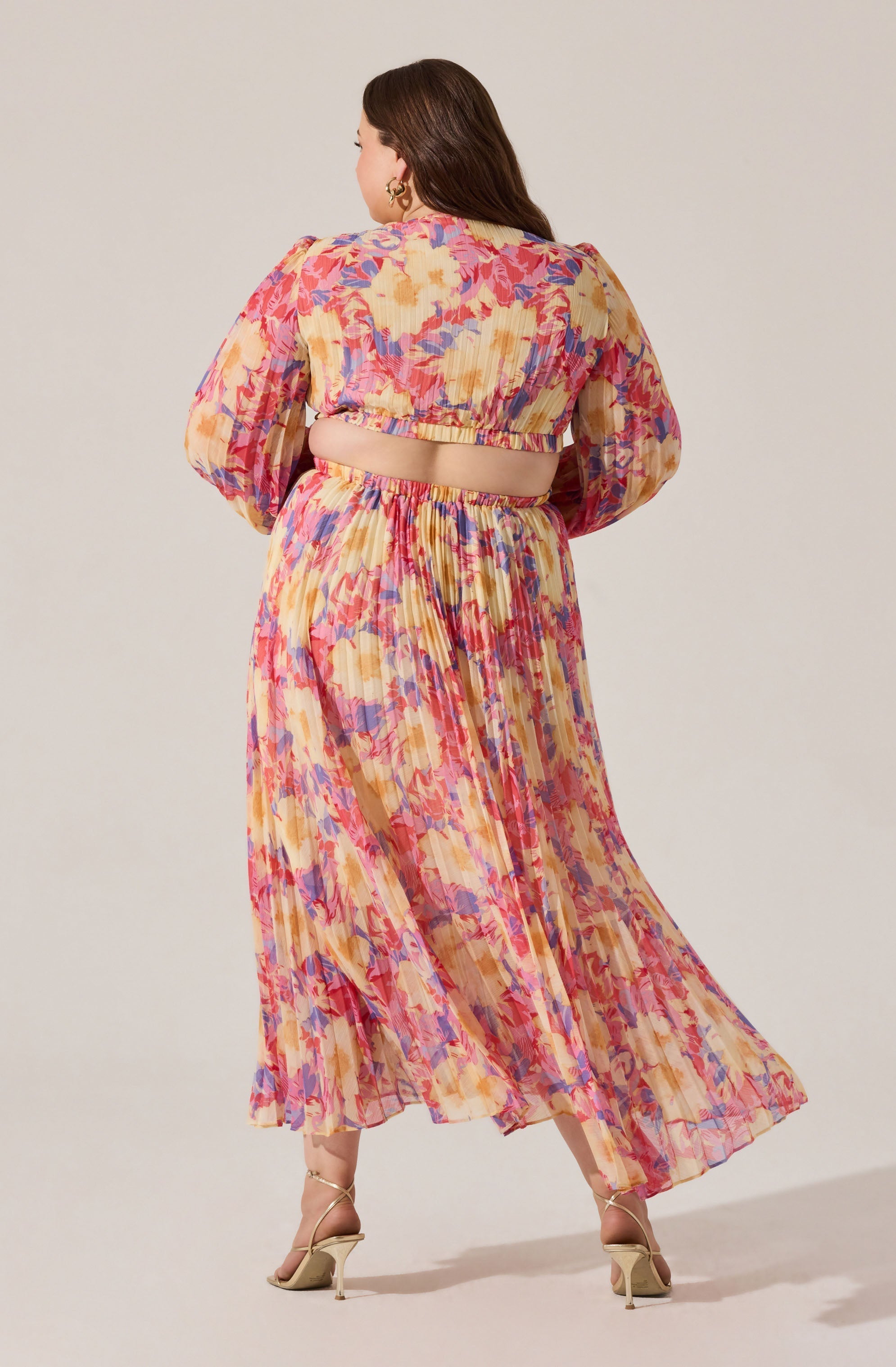 A woman is wearing a two-piece outfit with a colorful, floral pattern. The top features long, puffed sleeves and a cropped length, exposing the midriff. The skirt is a high-waisted, ankle-length, flowy maxi with a matching vibrant, abstract floral print. The fabric appears lightweight and semi-sheer with pleats. She pairs the outfit with nude high-heeled sandals.