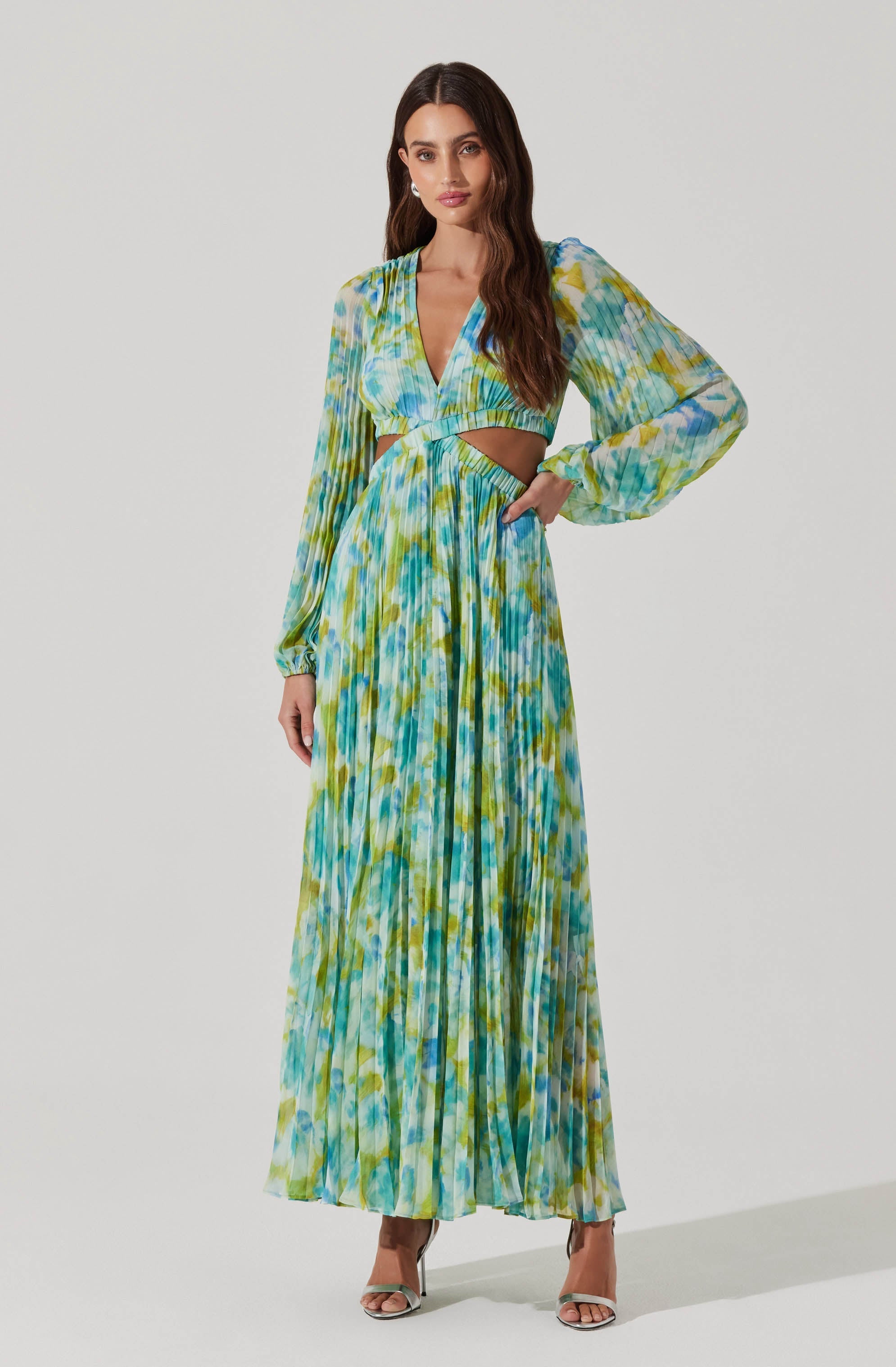 The dress features a vibrant, tie-dye pattern with shades of blue, green, yellow, and white. It has long, slightly puffed sleeves with elastic cuffs, a deep V-neckline, and a cut-out detail at the waist. The skirt is long and flowing, with pleats that add texture and movement. The overall look is colorful and bohemian.
