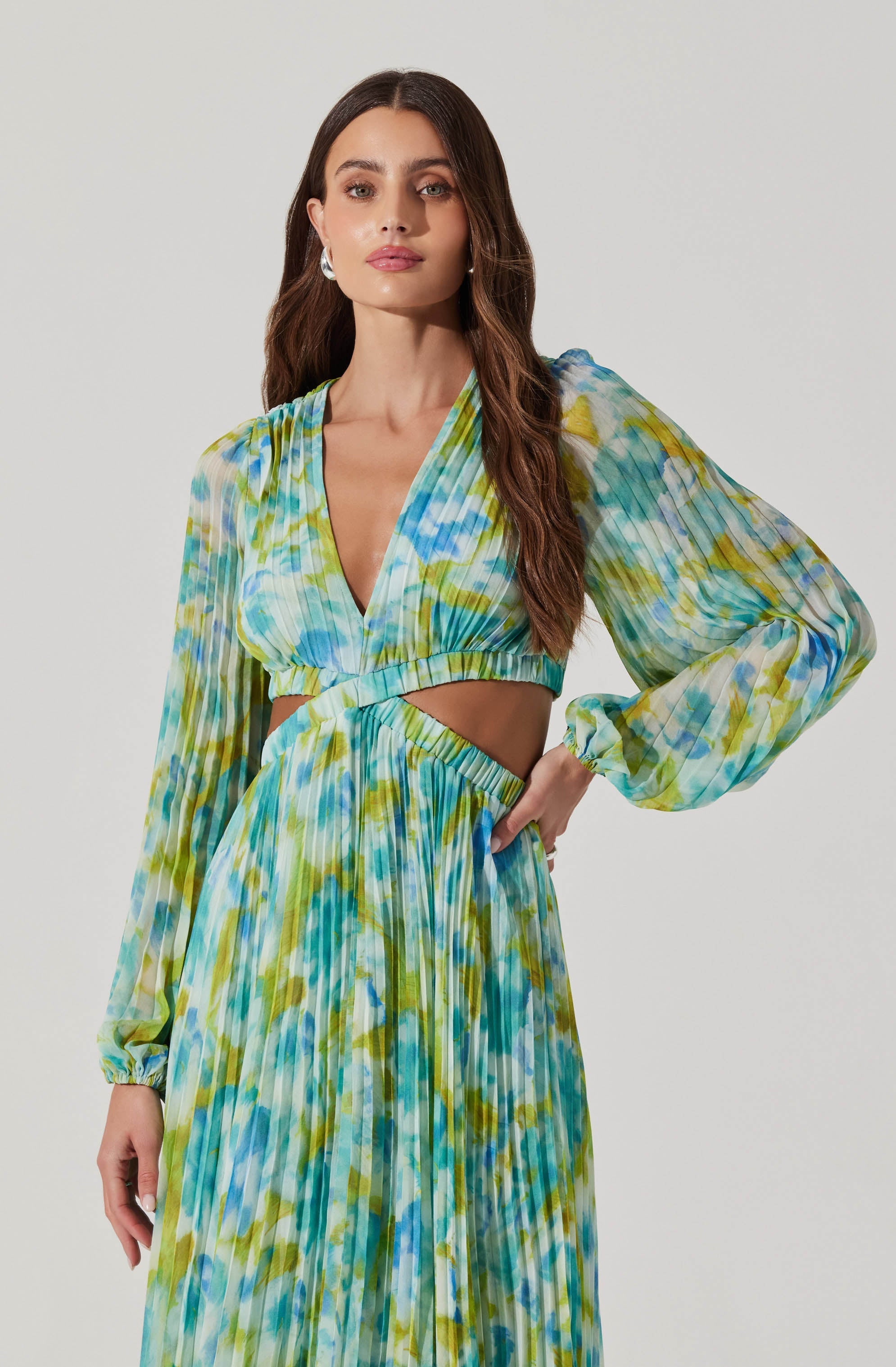 A woman is wearing a two-piece outfit with a vibrant, multicolored, watercolor-like pattern in shades of blue, green, and yellow. The top has long, semi-sheer, puffed sleeves with elastic cuffs, a V-neckline, and a cut-out detail at the midriff. The skirt is high-waisted, flowing, and pleated, matching the pattern of the top. The overall look is stylish and summery.