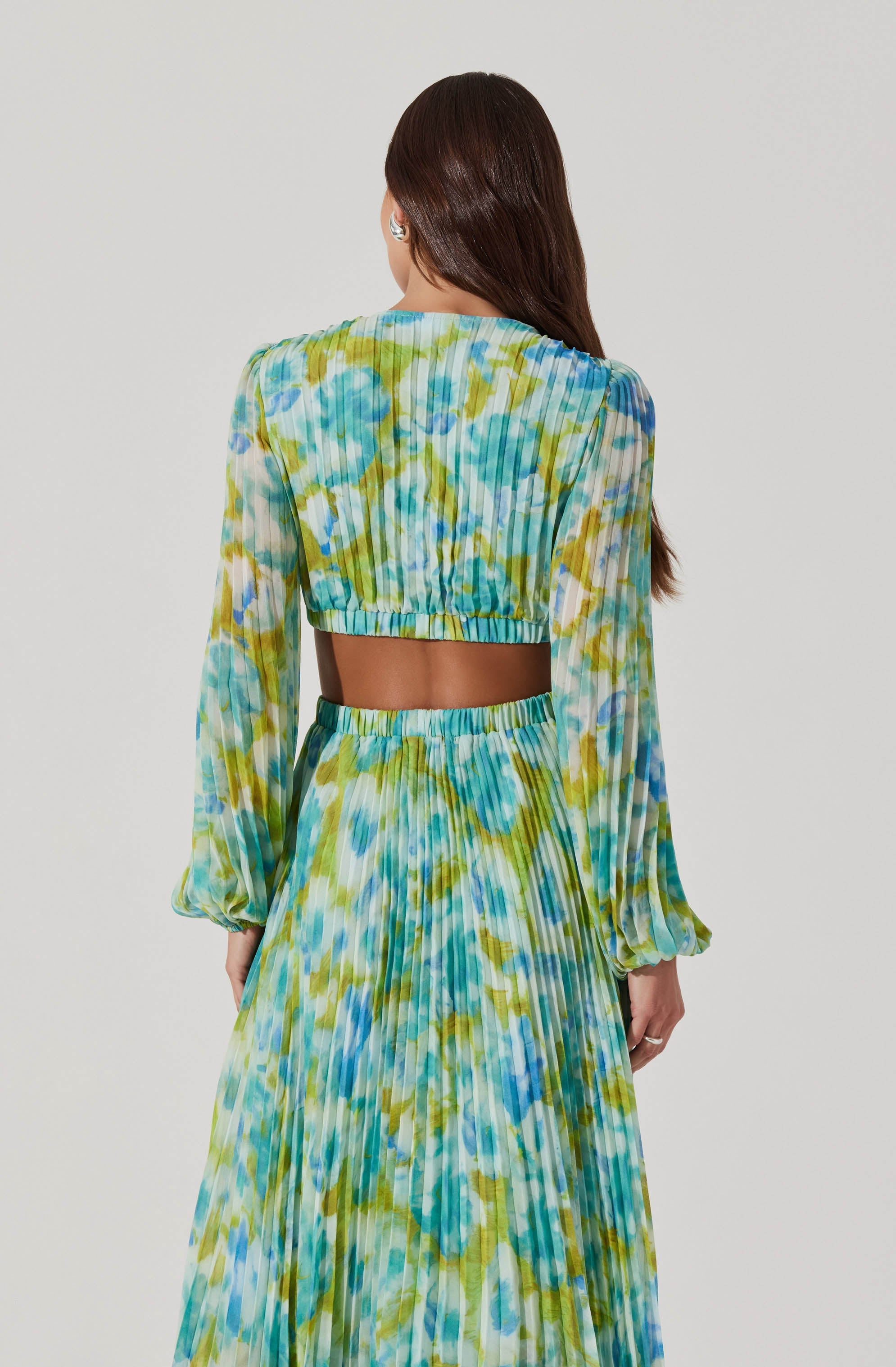 A woman is wearing a two-piece matching outfit with a cropped long-sleeve top and a high-waisted, pleated maxi skirt. The outfit features a vibrant, abstract tie-dye pattern in shades of blue, green, and yellow. The top has a rounded neckline and elasticized cuffs on the sleeves.
