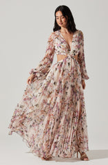 A woman is wearing a two-piece floral outfit with a deep V-neck crop top that ties at the front, featuring long, semi-sheer, puffed sleeves with elastic cuffs. The matching high-waisted, pleated maxi skirt has a flowing, semi-sheer fabric with a multicolored floral pattern. She is also wearing strappy high-heeled sandals.