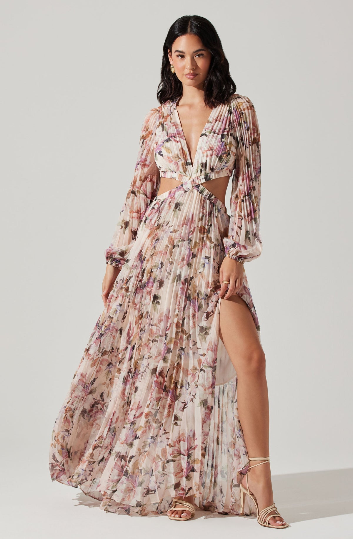 A woman is wearing a long, flowing dress with a floral pattern in soft pink, beige, and purple tones. The dress has long, sheer sleeves with elastic cuffs, a deep V-neckline, and cut-out sections at the waist that reveal her midriff. The skirt of the dress is high slit, showing her leg. She is also wearing strappy, beige high-heeled sandals.