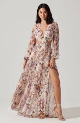 A woman is wearing a long, flowing dress with a floral pattern in soft pink, beige, and purple tones. The dress has long, sheer sleeves with elastic cuffs, a deep V-neckline, and cut-out sections at the waist that reveal her midriff. The skirt of the dress is high slit, showing her leg. She is also wearing strappy, beige high-heeled sandals.