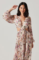 A woman is wearing a long, flowy dress with a floral print in shades of pink, purple, and green. The dress features a deep V-neckline, long puffed sleeves, and cut-out sections at the waist, with a gathered waistline that flows into a pleated skirt.