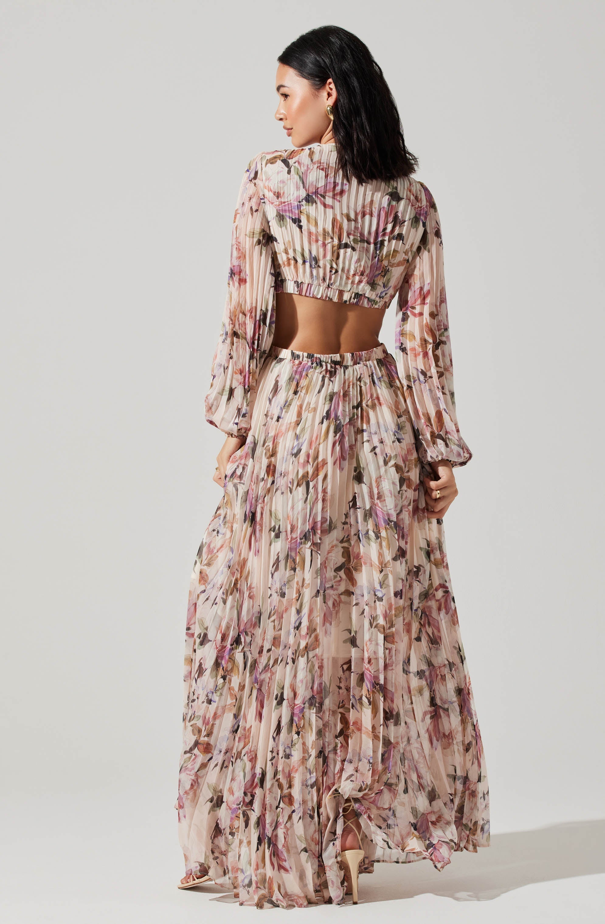 A woman is wearing a matching two-piece dress set with a floral pattern in shades of pink, purple, and brown. The top is a long-sleeved, cropped blouse with a smocked waistband and slightly puffed sleeves. The skirt is a high-waisted, flowing, full-length gown with a pleated design and an asymmetrical hemline. The fabric appears lightweight and semi-sheer with a soft, dreamy floral print. She is also wearing open-toe high heels.