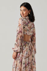 A woman with shoulder-length dark hair is wearing a matching floral outfit consisting of a long-sleeved, cropped top and high-waisted, flowing pants. The top features a round neckline, puffed sleeves, and an elastic band at the hem. The fabric of both pieces is sheer with a pleated texture, adorned with a floral pattern in shades of pink, purple, brown, and green. She is accessorized with large, heart-shaped gold earrings.
