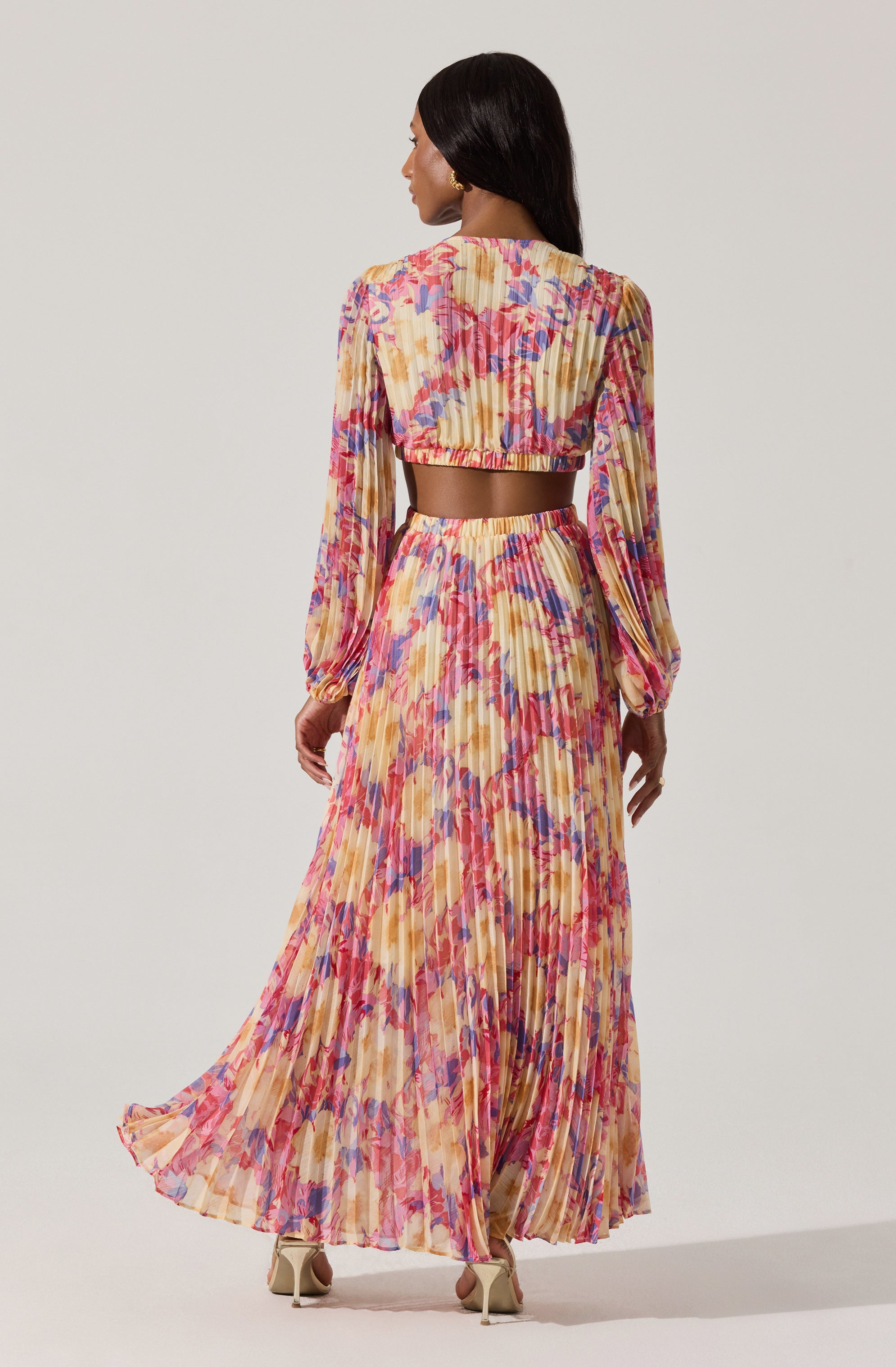 A woman is wearing a two-piece outfit with a colorful, floral, pleated design. The top is a cropped, long-sleeve blouse with puffed sleeves and a ruffled, elasticized neckline. The matching skirt is high-waisted, floor-length, pleated, and features a vibrant pattern of brushstroke-style flowers in shades of pink, purple, yellow, and orange. She is also wearing beige high-heeled shoes.