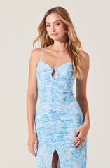A woman is wearing a fitted, sleeveless dress with thin spaghetti straps. The dress features a light blue and white watercolor-inspired pattern, ruched detailing along the front, and a small keyhole cutout at the neckline. The bottom of the dress has a front slit.